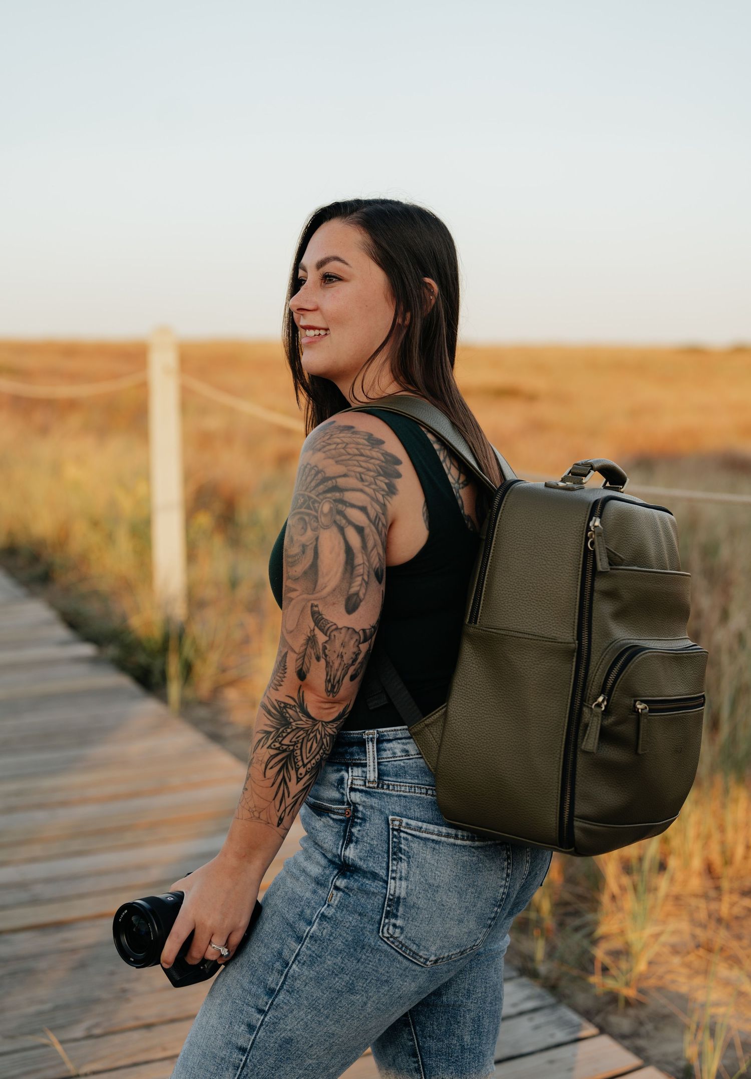 Woman with tattoos wearing jeans and olive backpack poses in golden field at sunset.