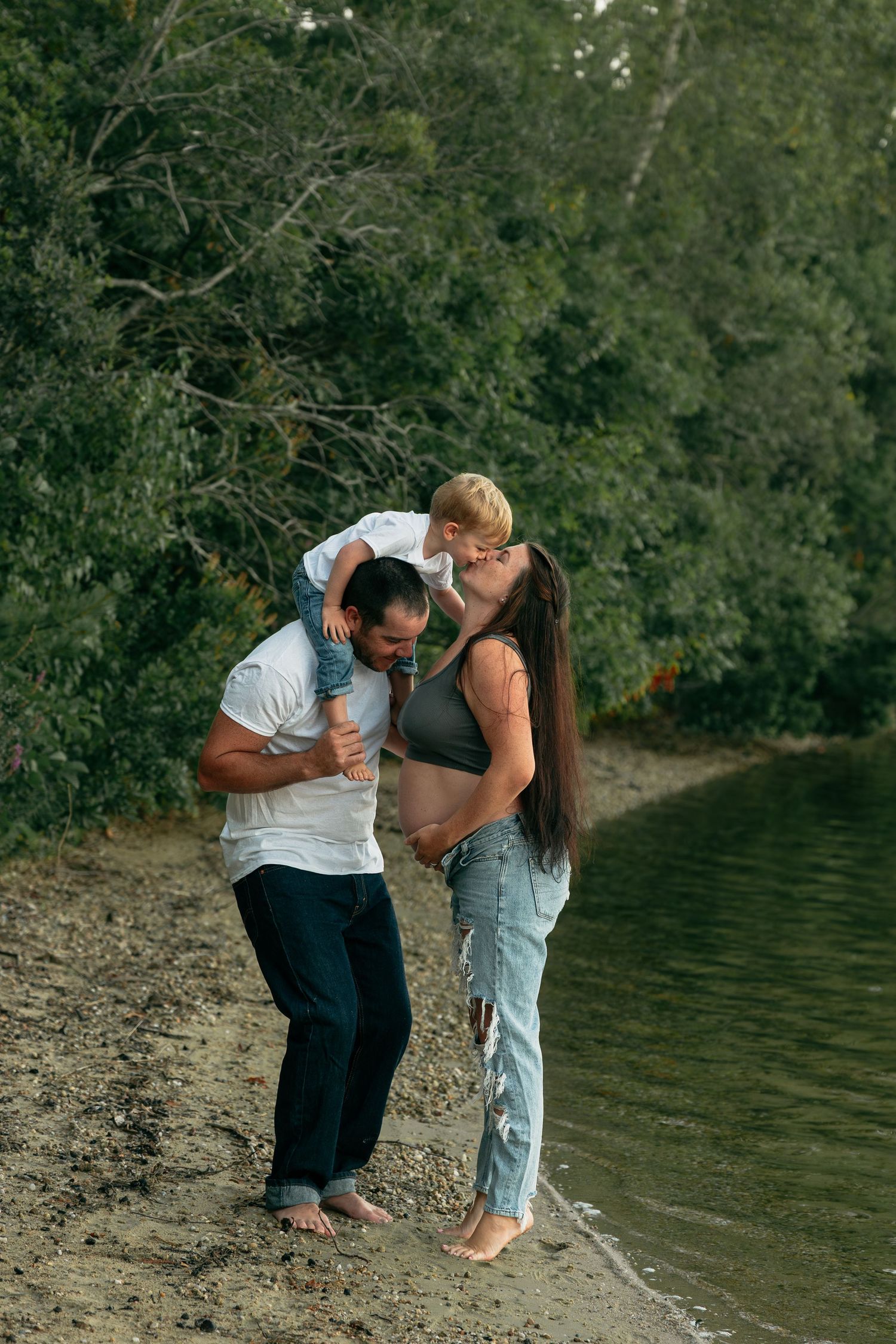 A loving family moment captured on a peaceful lakeside beach with green foliage in background.