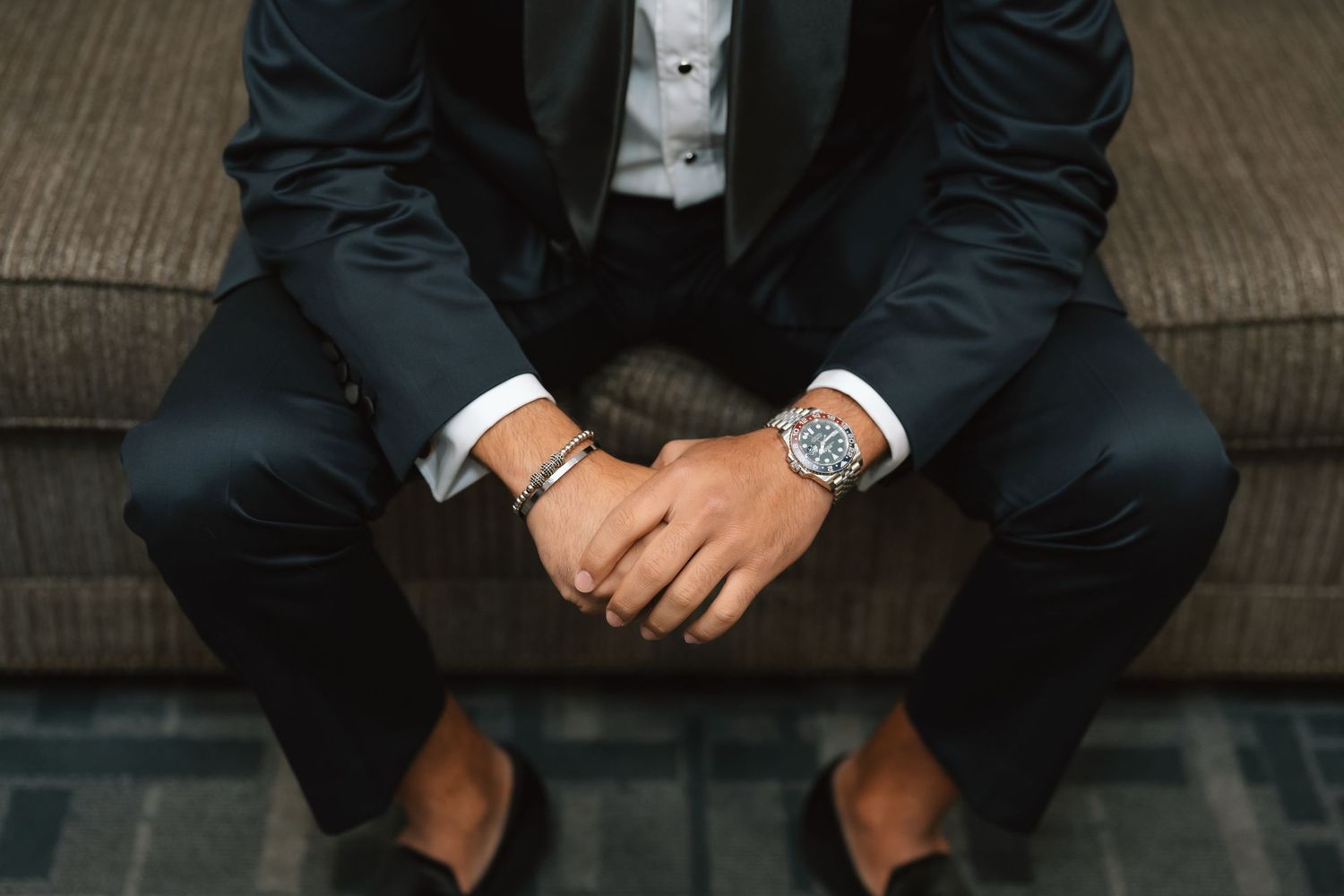 Detail shot of groom's hands with watch sitting on dark couch in formal attire.