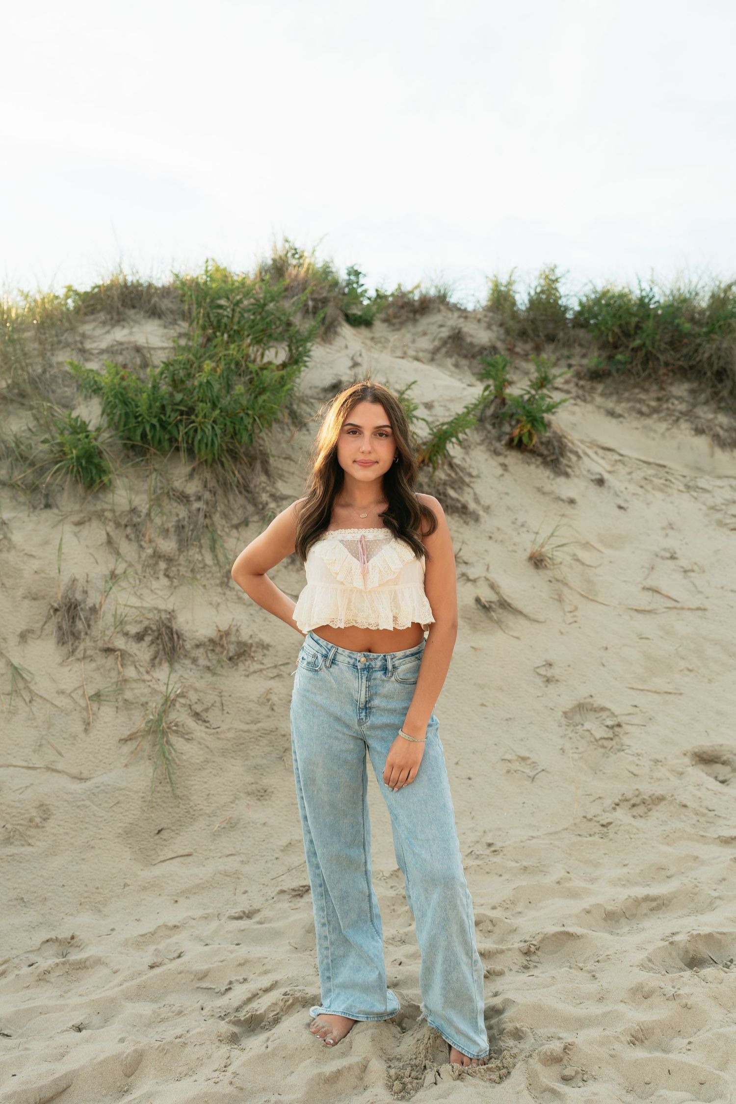 A person in casual denim and white top poses on sandy dunes with beach grass.