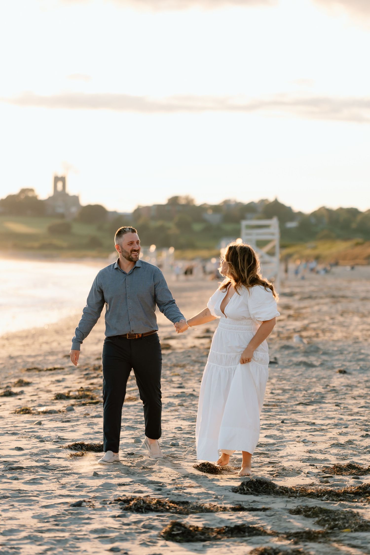 A couple walks together on a beach with a lighthouse visible in the background.