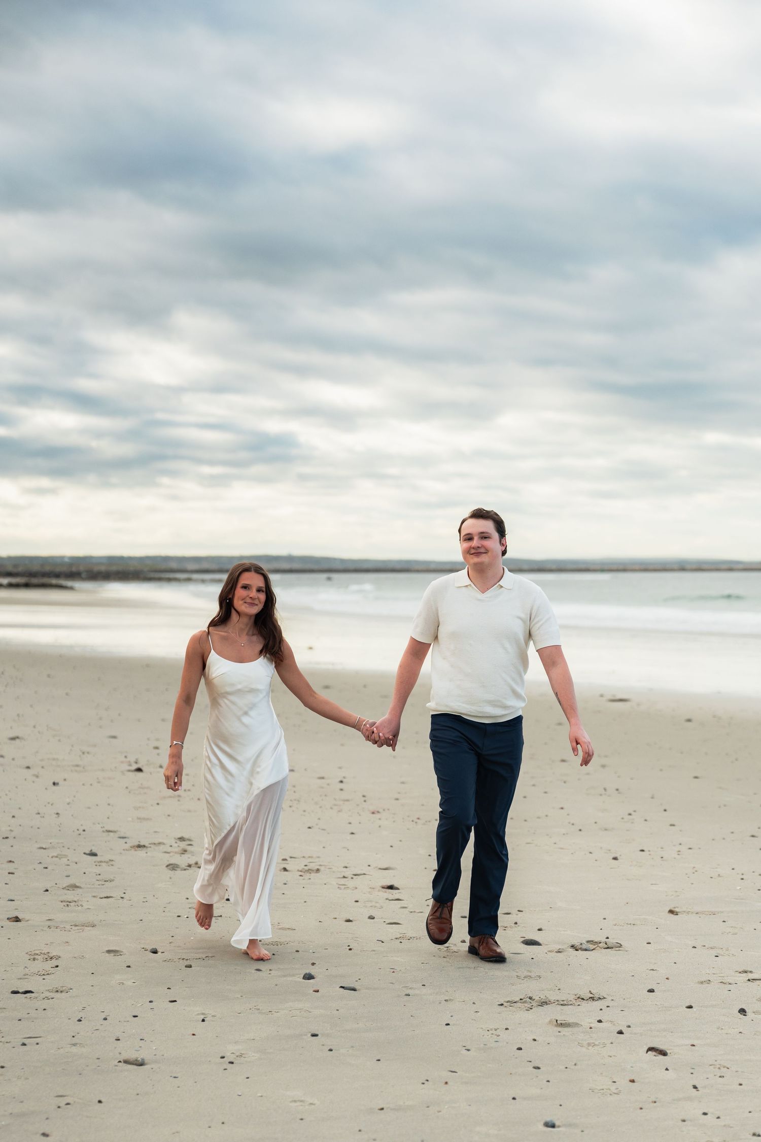 A couple running playfully together on an empty beach under cloudy skies.
