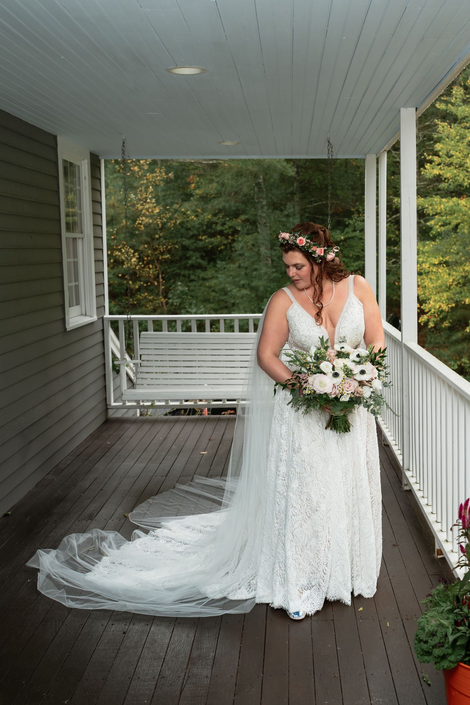 A bride in a lace wedding dress with floral crown stands on a covered porch holding a white bouquet.
