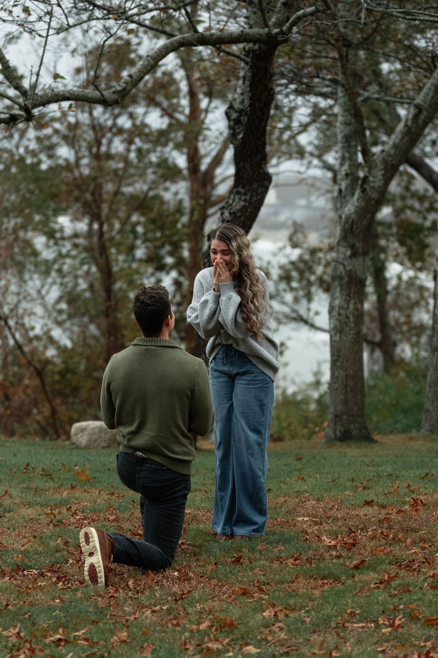 Couple standing in fallen autumn leaves with bare trees in background.