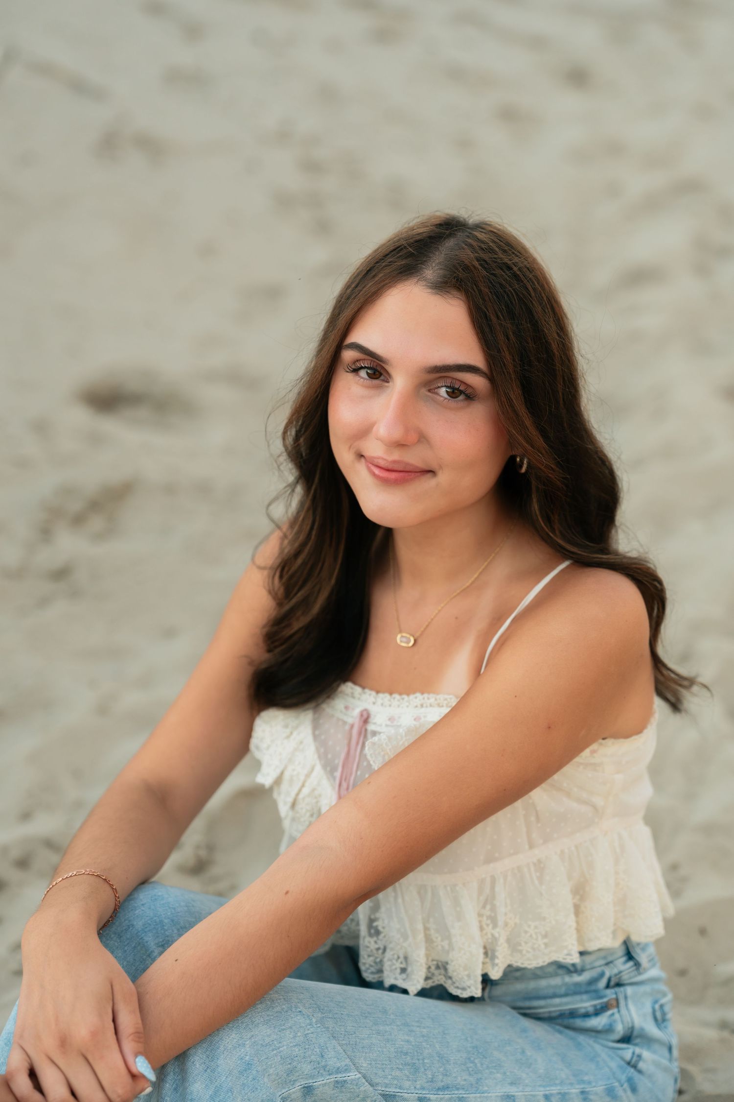 A person in jeans and white lace top poses on sandy beach during sunset.