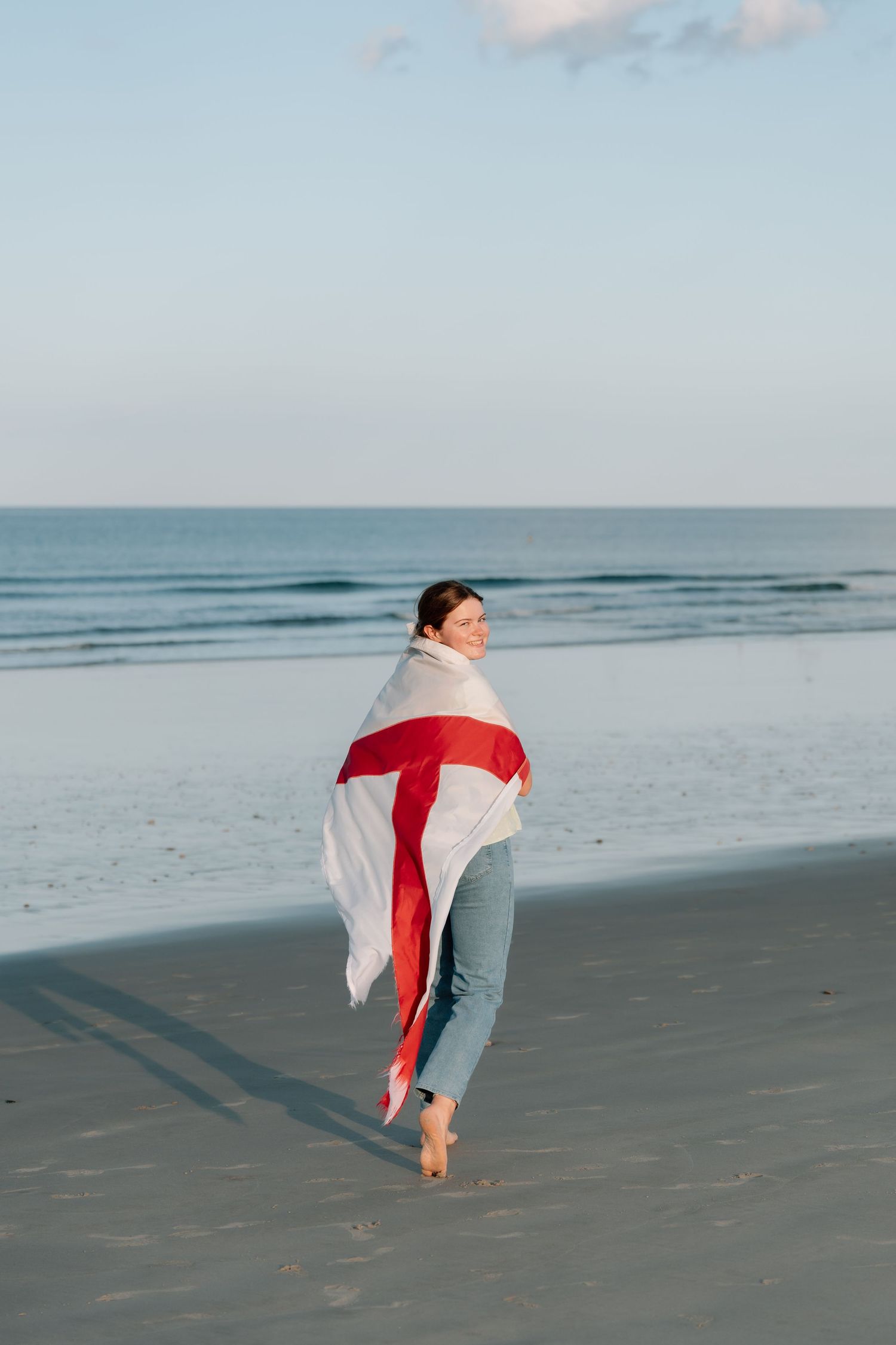 Figure wrapped in red and white fabric walking along empty beach at sunset.