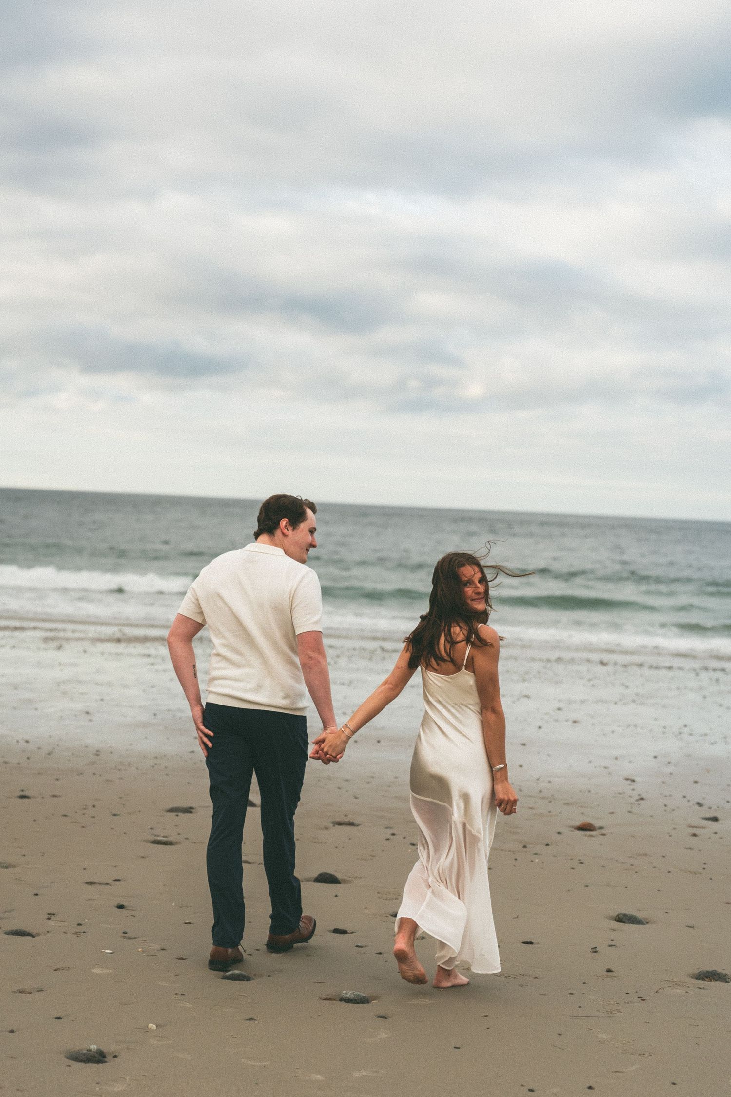 A couple playfully walks along a misty beach shore with crashing waves in the background.