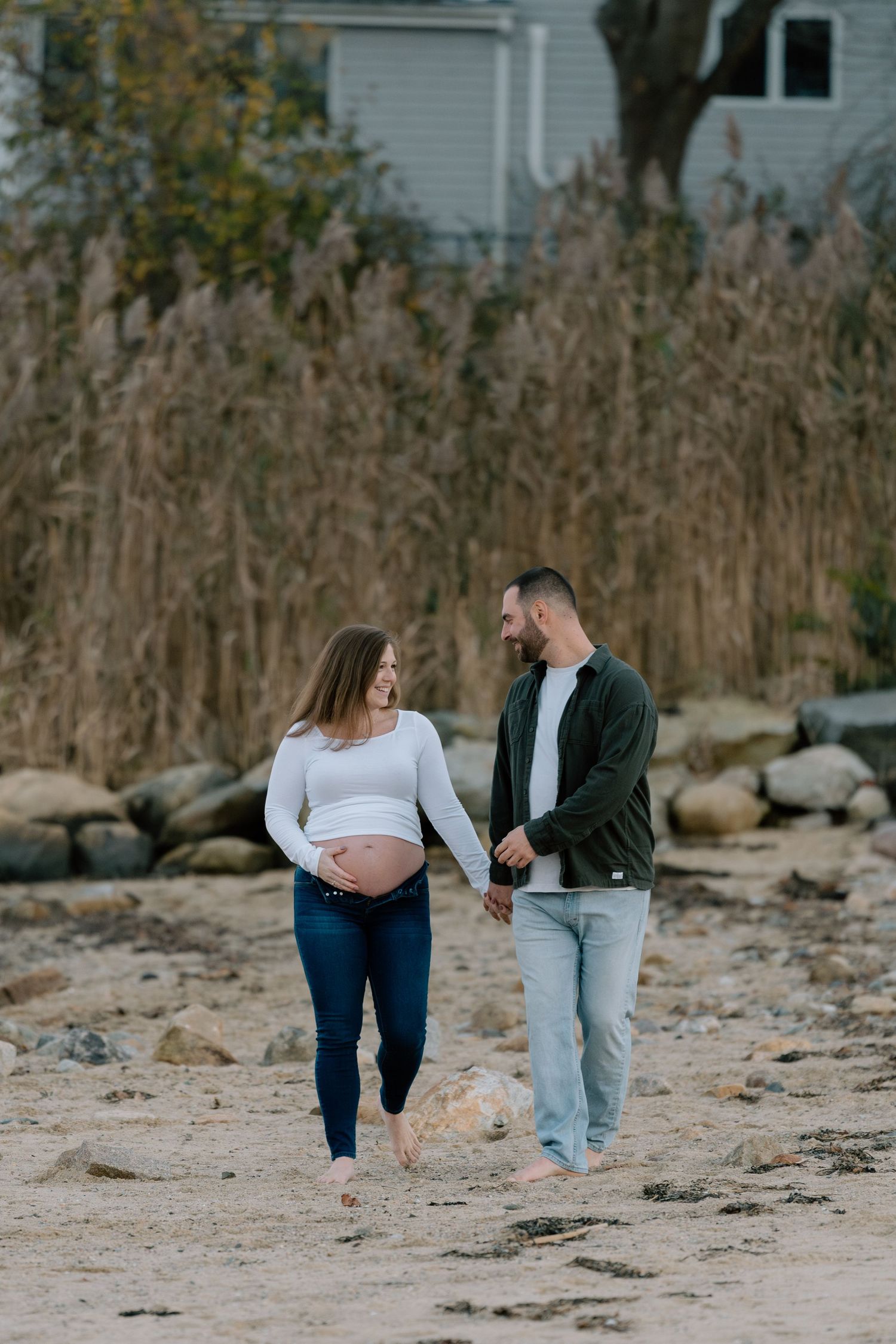 Couple walking along rocky beach shore during maternity photoshoot.