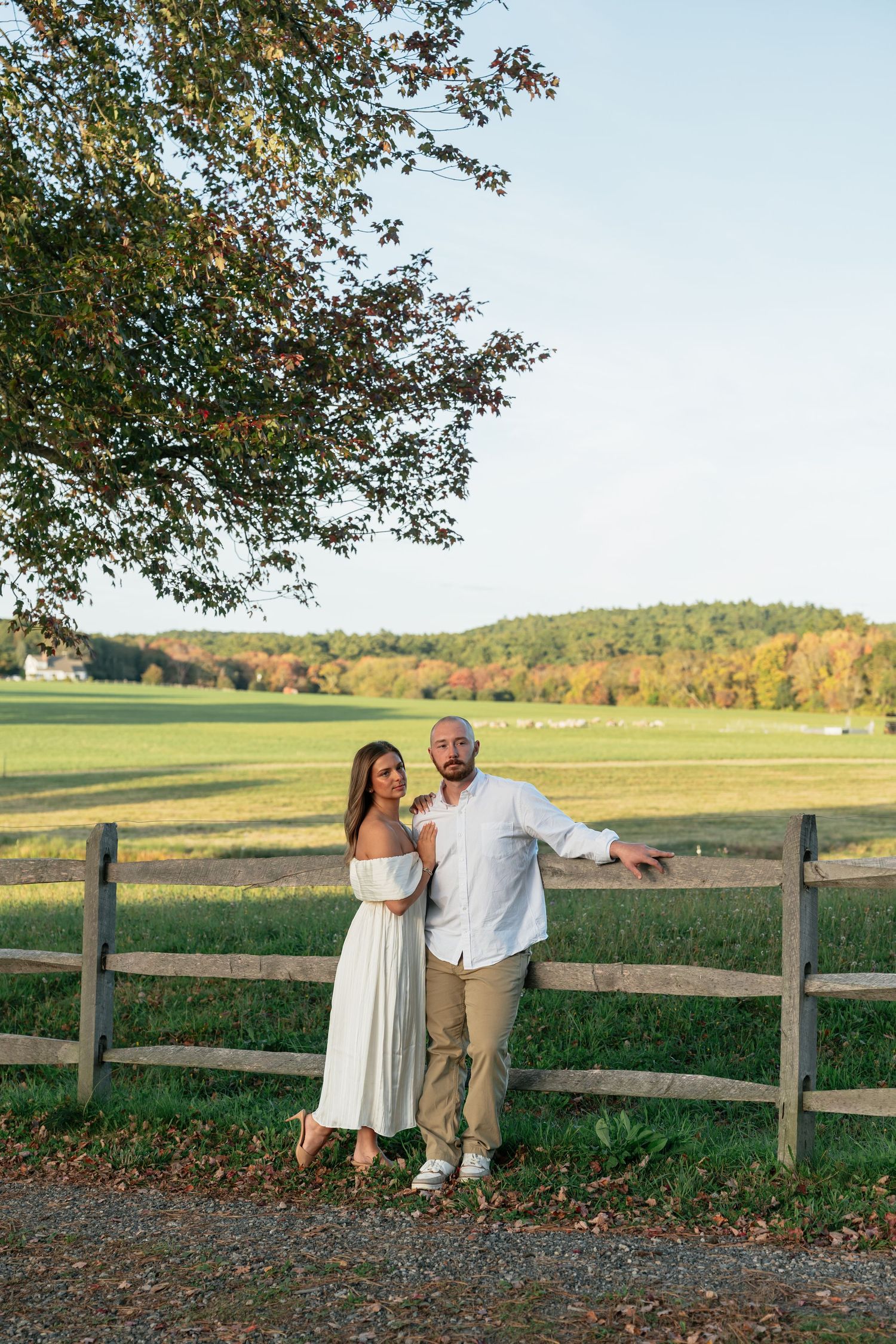 Couple stands by rustic wooden fence overlooking autumn countryside at sunset.