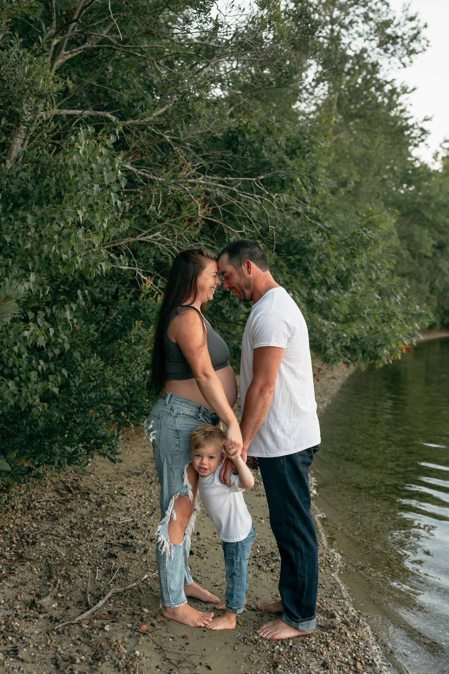A couple shares an intimate moment by the water's edge surrounded by trees.