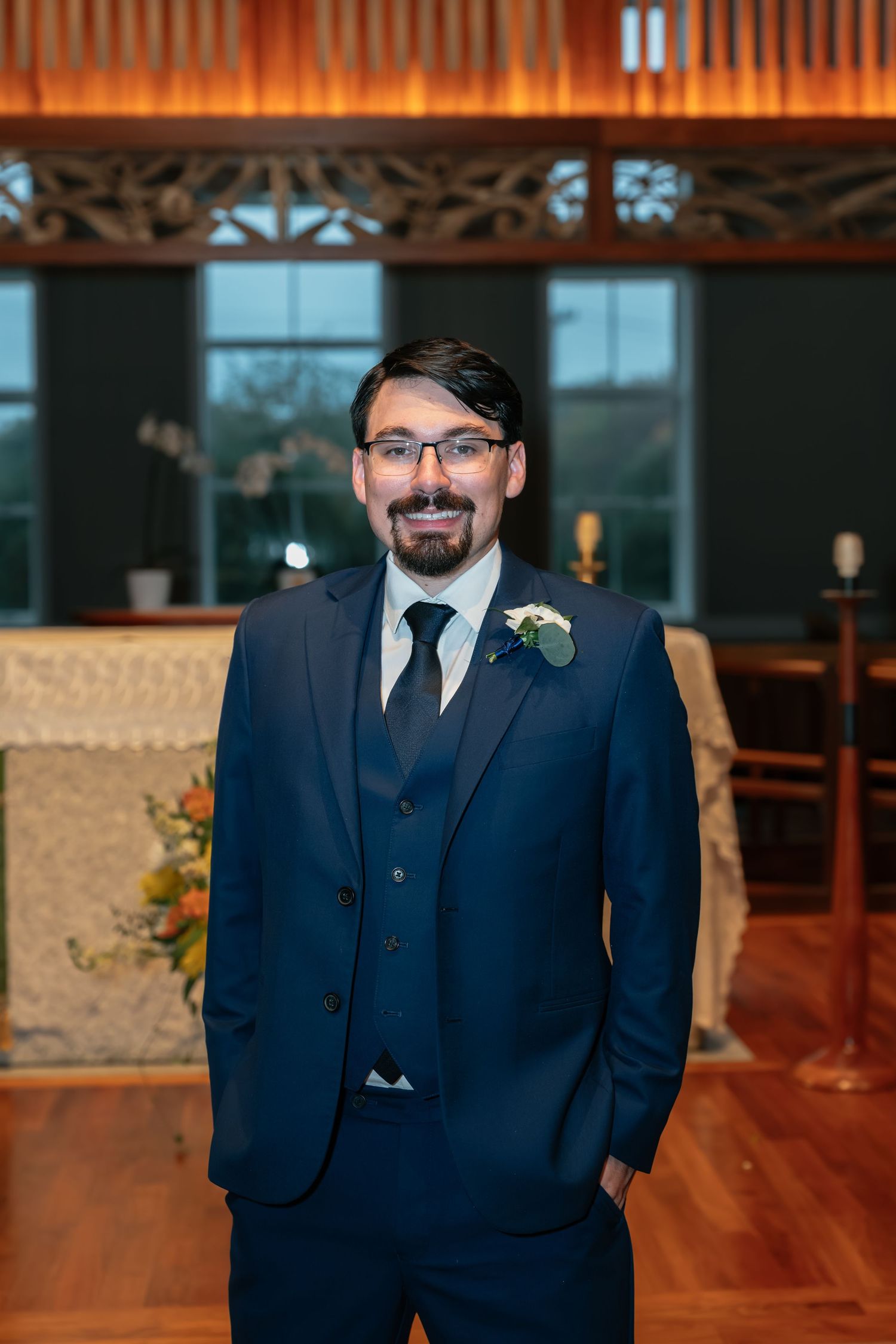 Groom in navy three-piece suit with boutonniere stands in church reception area.