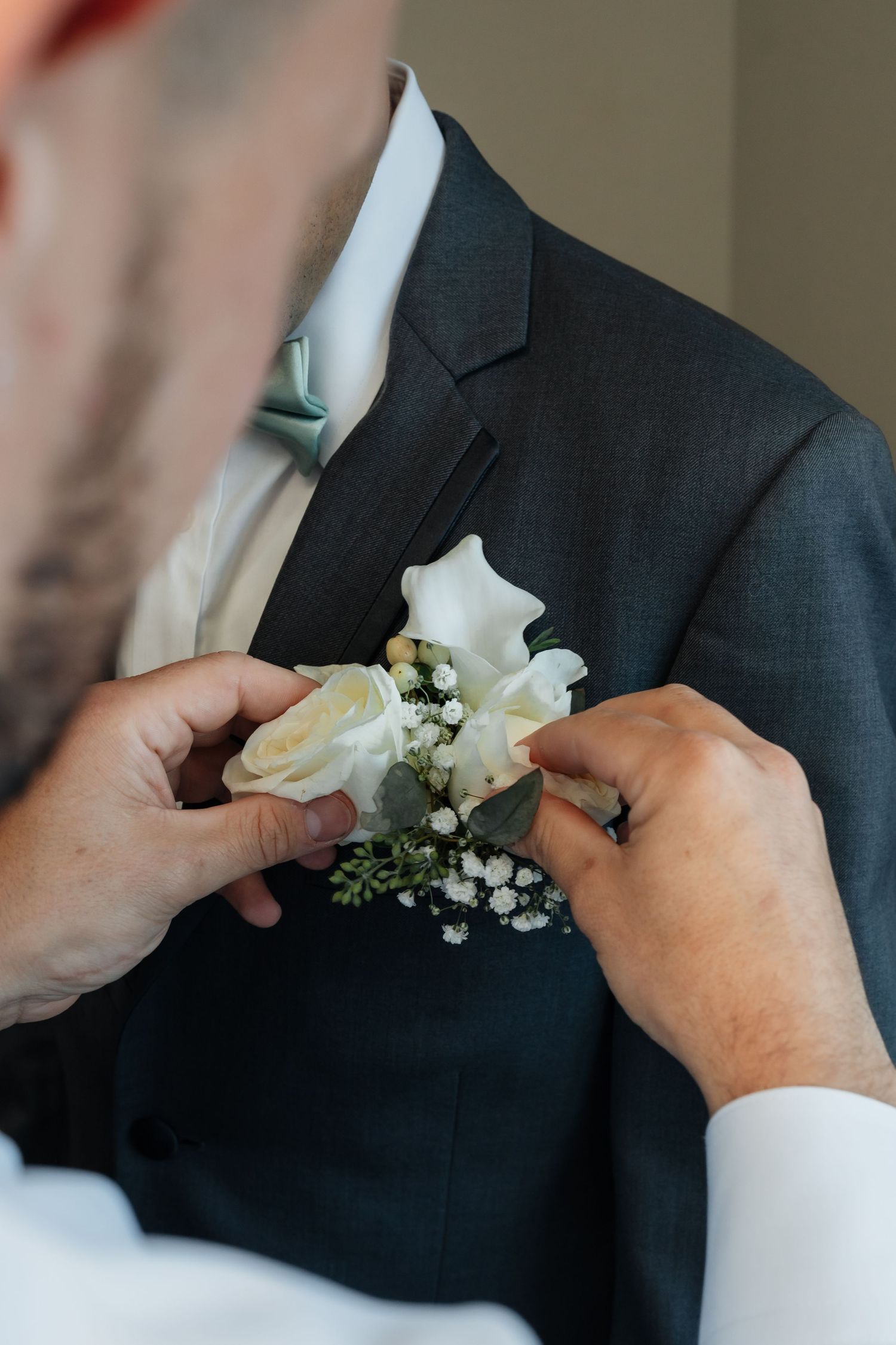 Hands pinning a white floral boutonniere onto a dark suit jacket.