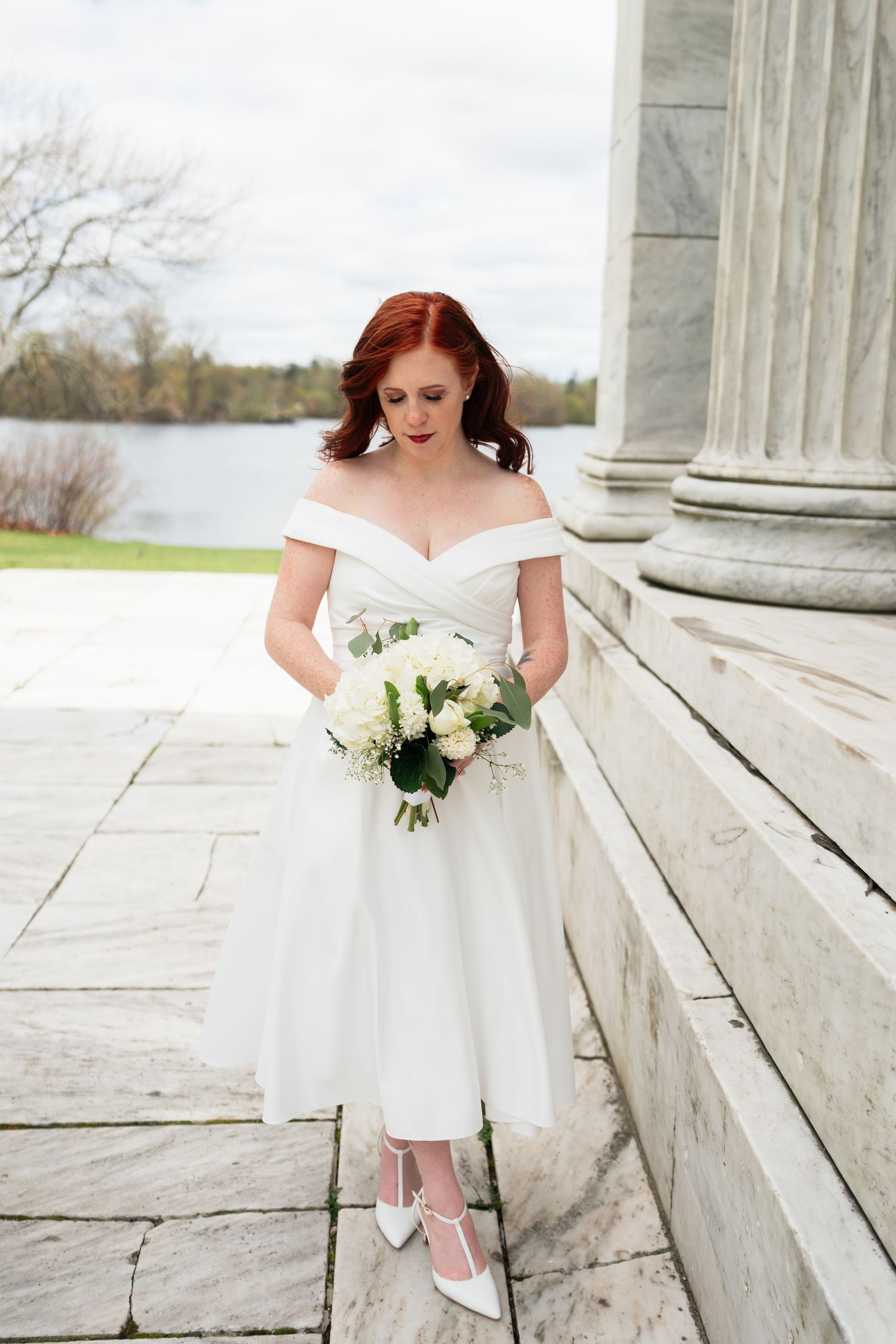 Bride in tea-length dress holding white bouquet by classical stone columns.