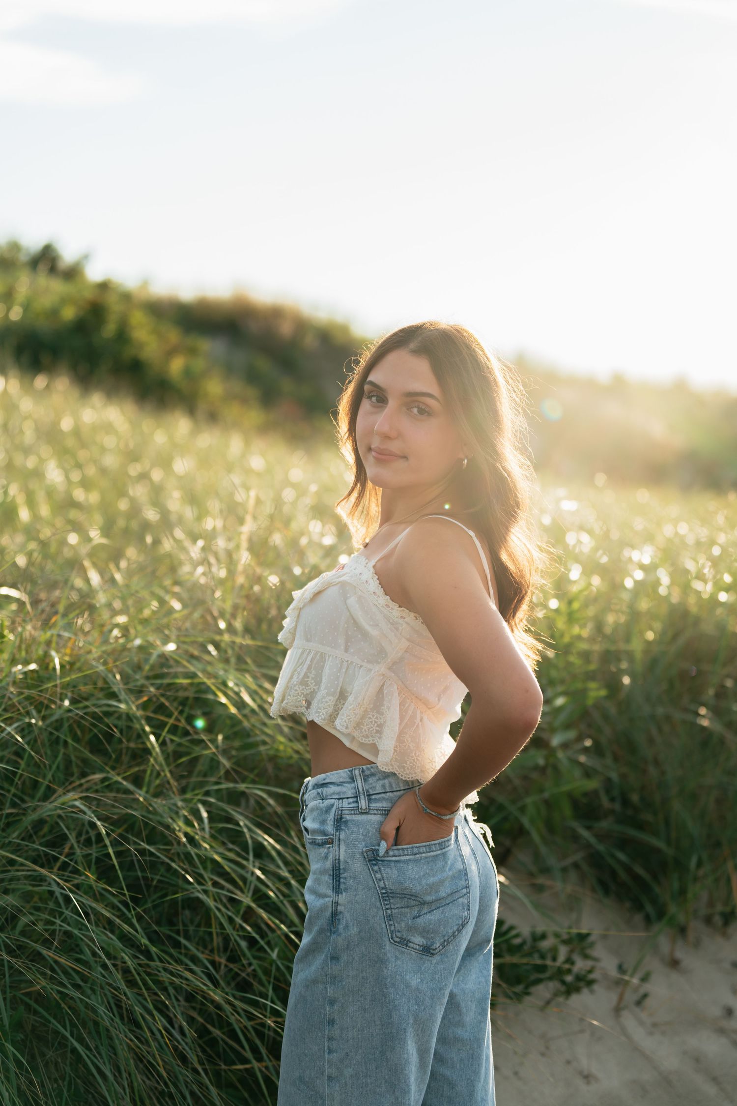 A person in denim and white top poses at sunset among beach grass.