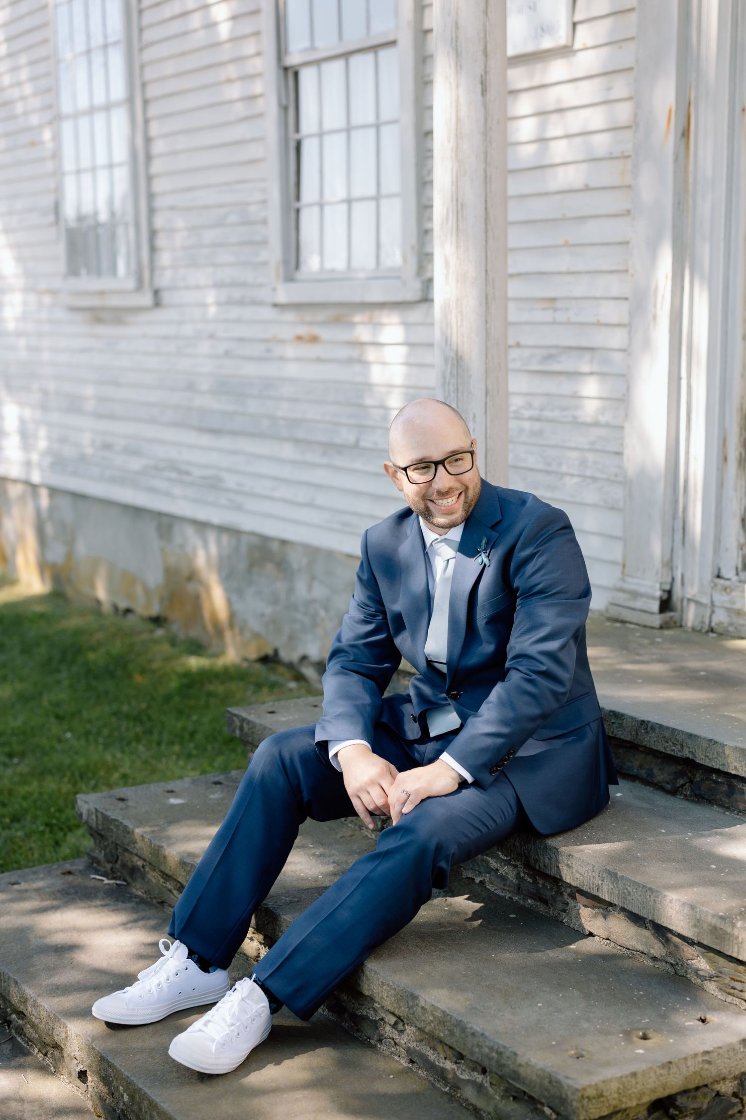A person in a navy suit and white sneakers sits casually on stone steps of a white building.