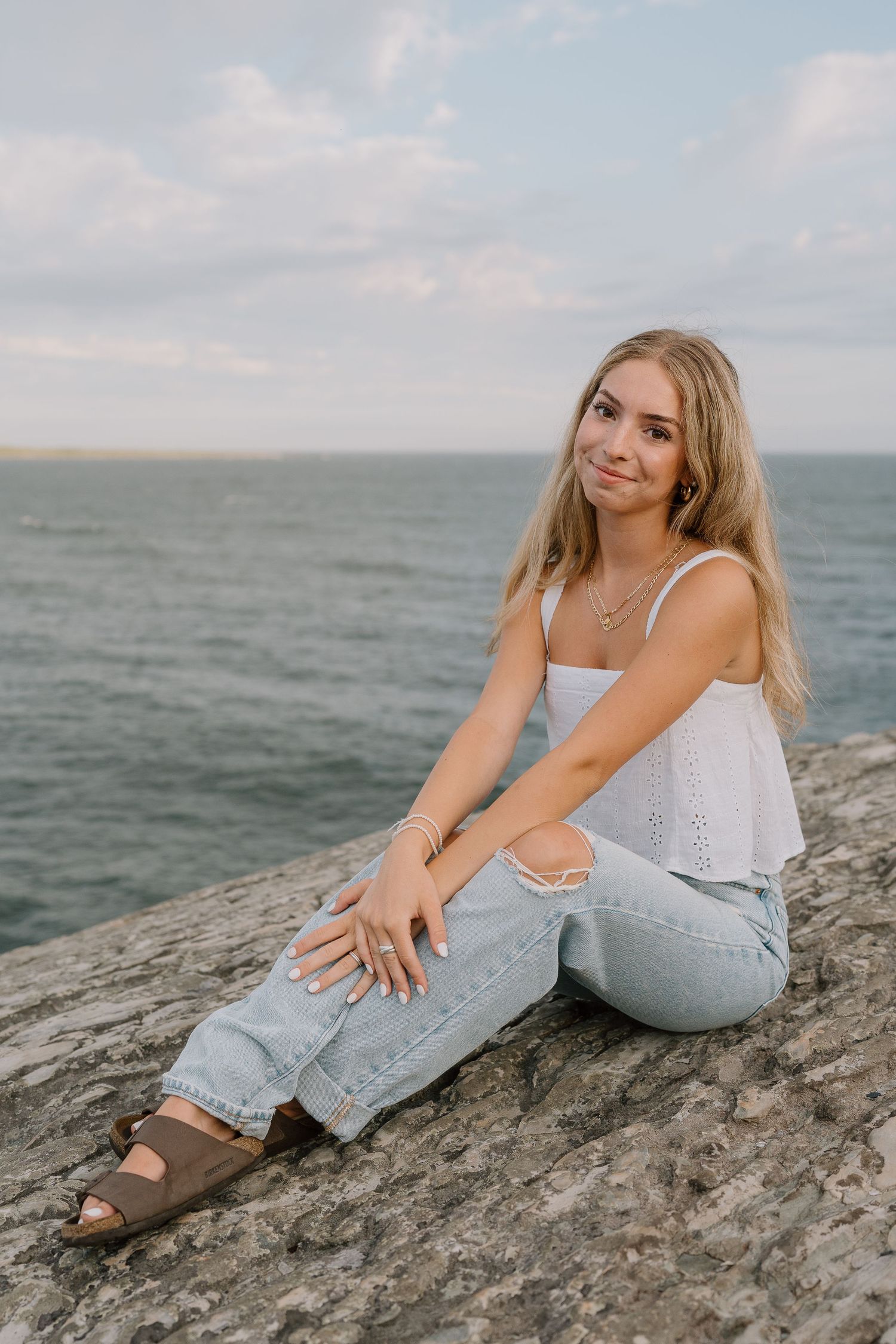 Relaxed pose sitting on coastal rocks wearing white tank top and light wash jeans.