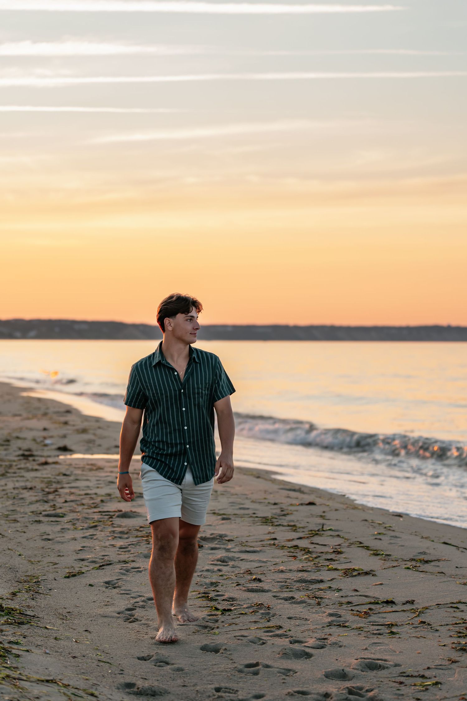 Person in striped shirt and white shorts walks along beach shoreline at sunset.