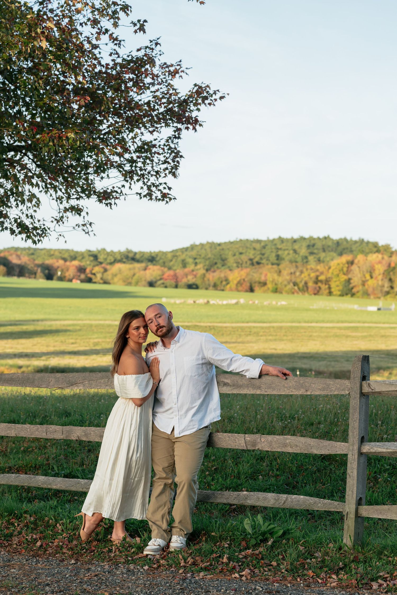 Romantic couple poses along countryside fence with fall foliage in background.