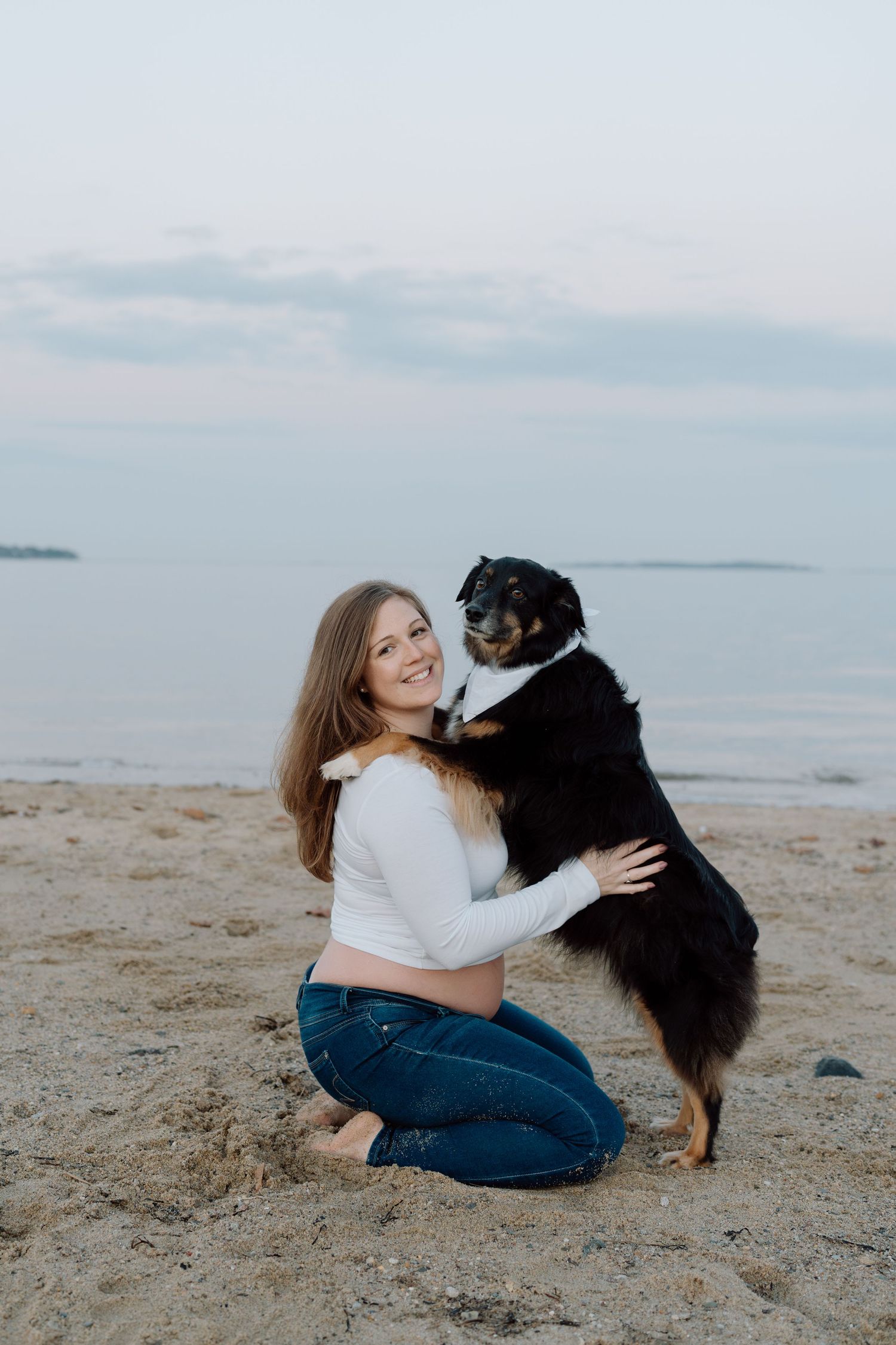 Expecting mother plays with black dog on sandy beach during sunset photoshoot.