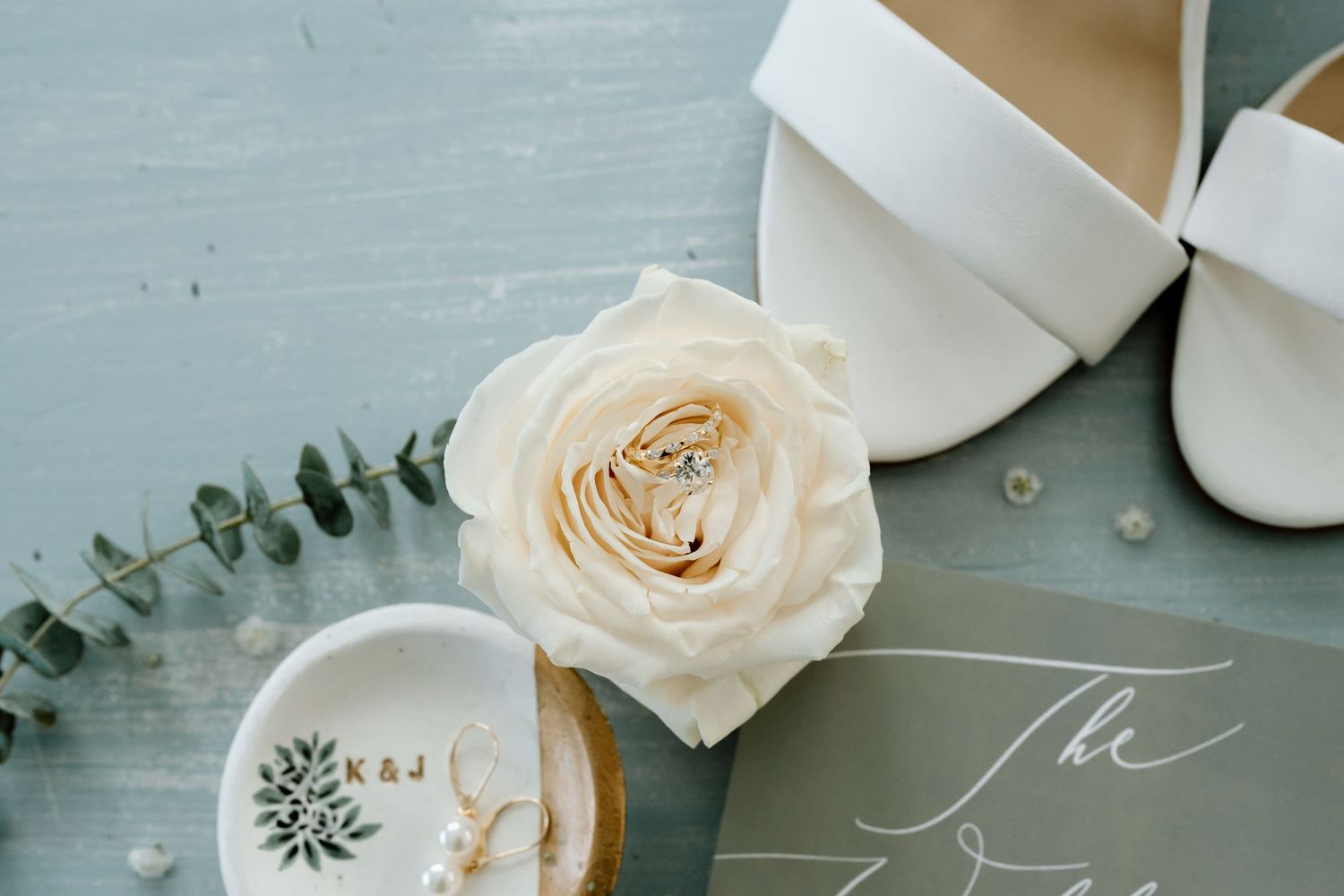 Cream rose and wedding accessories styled with eucalyptus on light blue background.