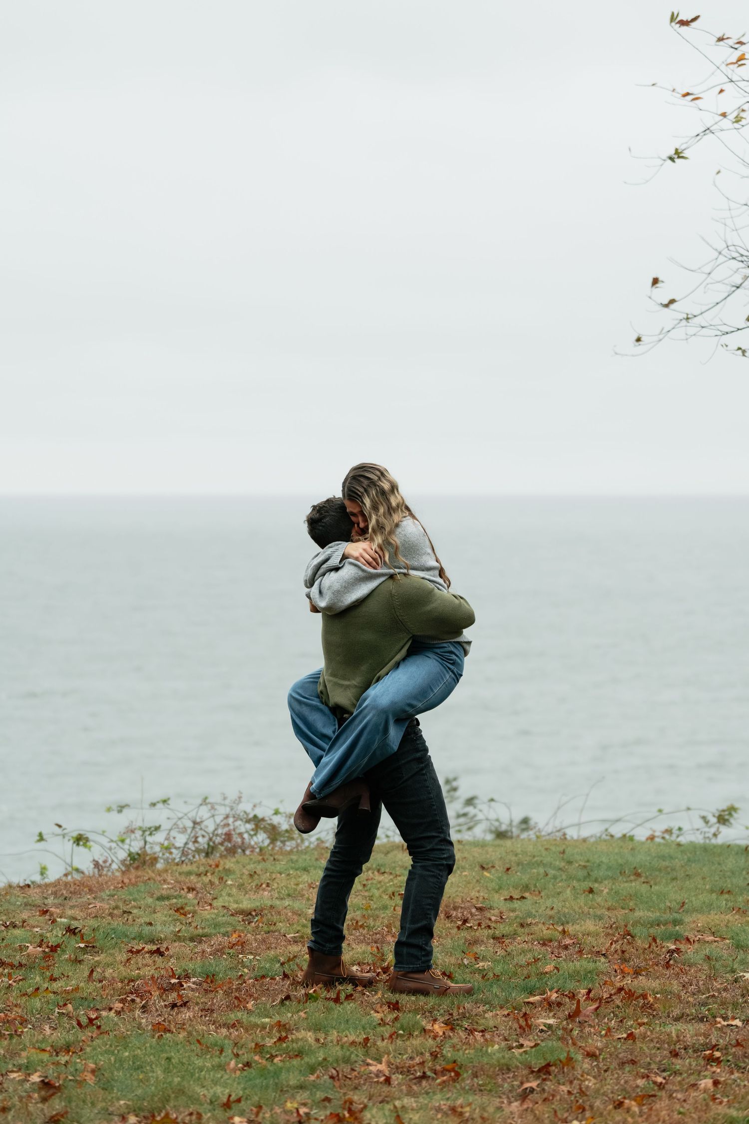 A couple embraces on a grassy cliff overlooking the ocean on an overcast autumn day.
