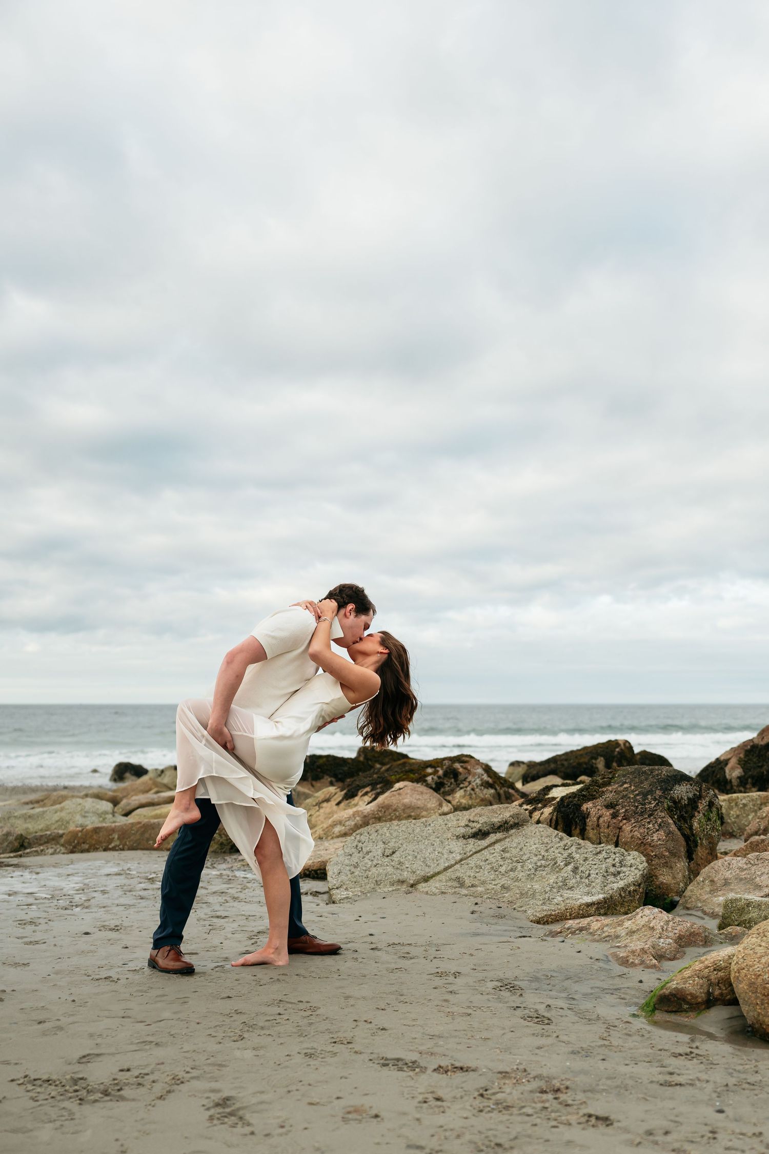 A romantic dip pose captured on a rocky beach with ocean waves in the background.