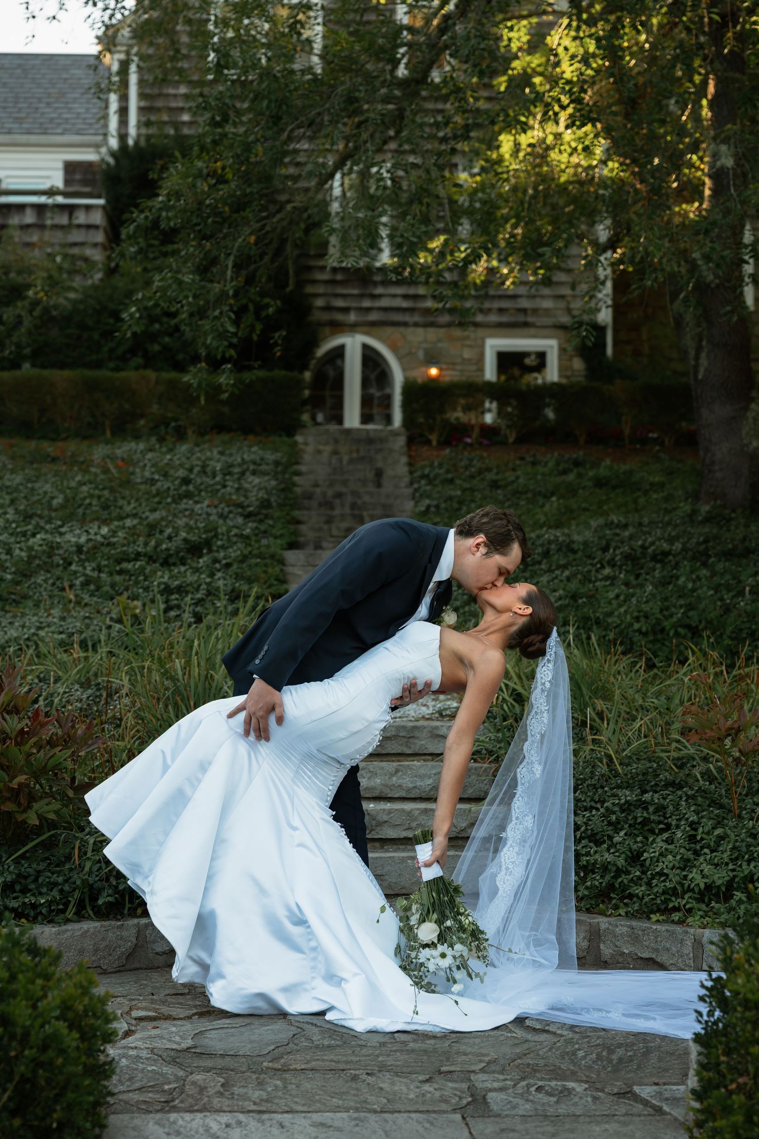 Wedding couple poses in a romantic dip on garden stone steps surrounded by greenery.