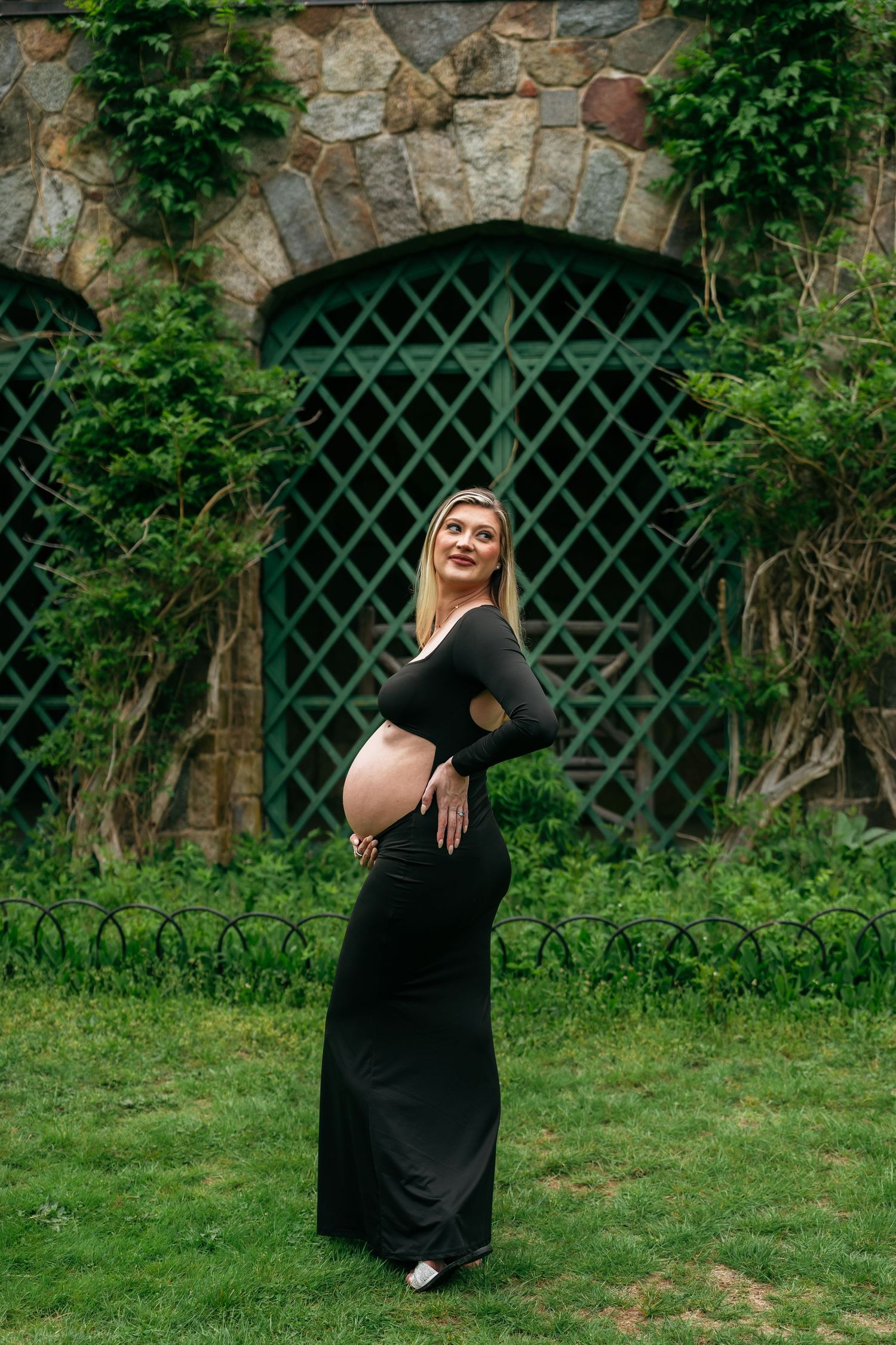 A maternity portrait session against a stone wall with green lattice backdrop in a garden setting.