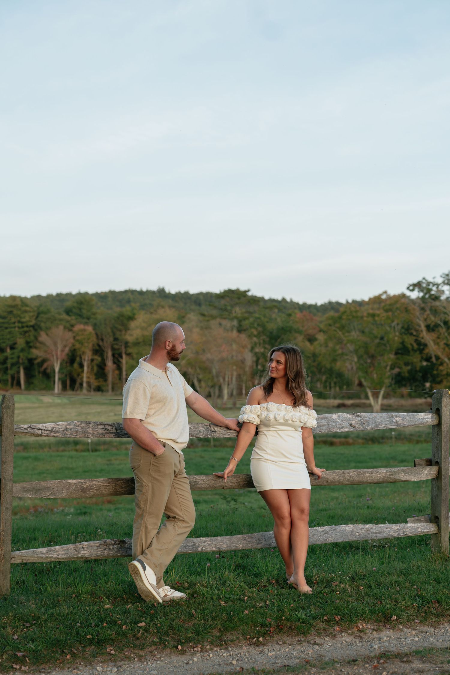 A couple playfully interacts along a wooden fence with rolling hills in the background.