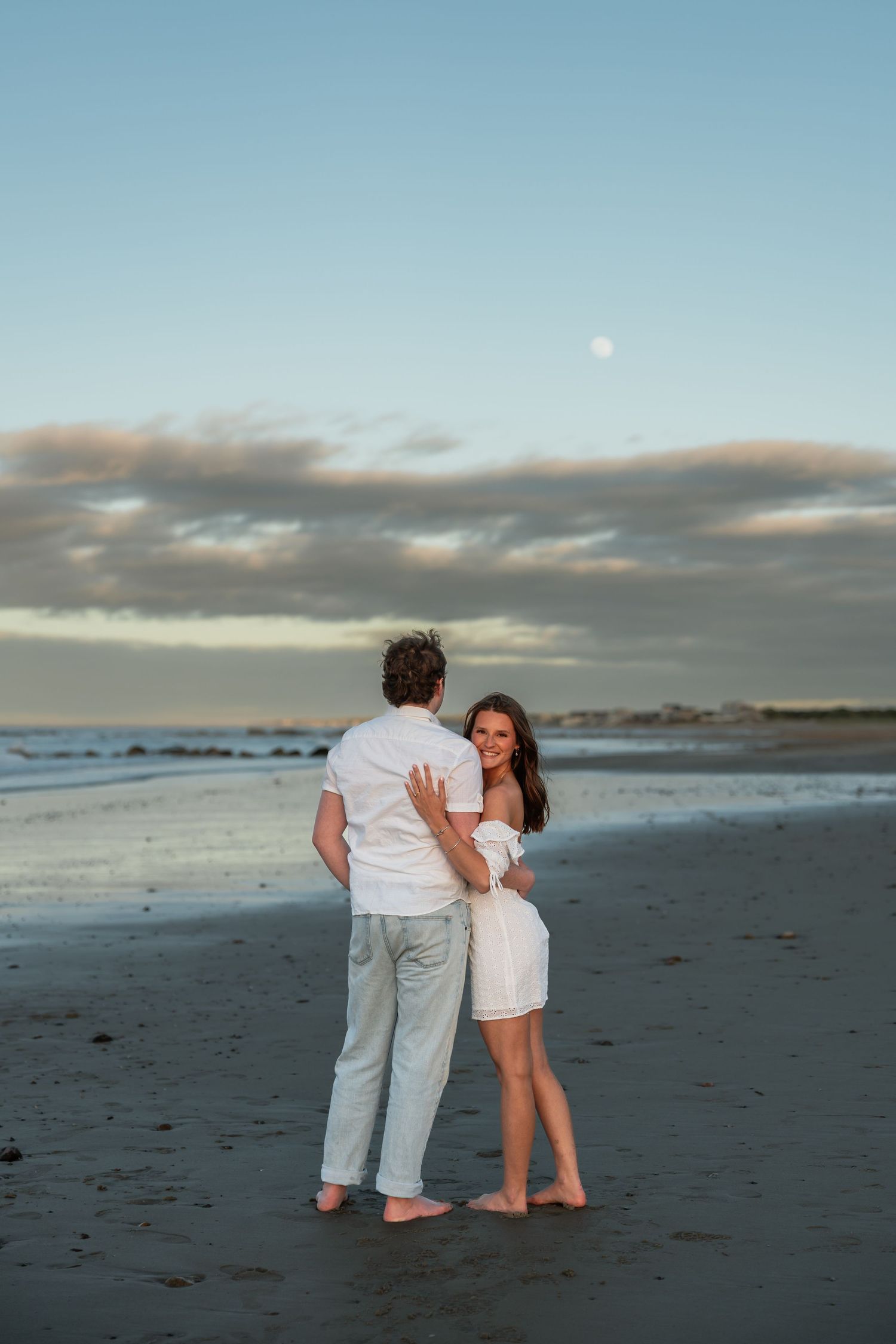 A couple shares playful moments on a beach with the moon visible in the evening sky.