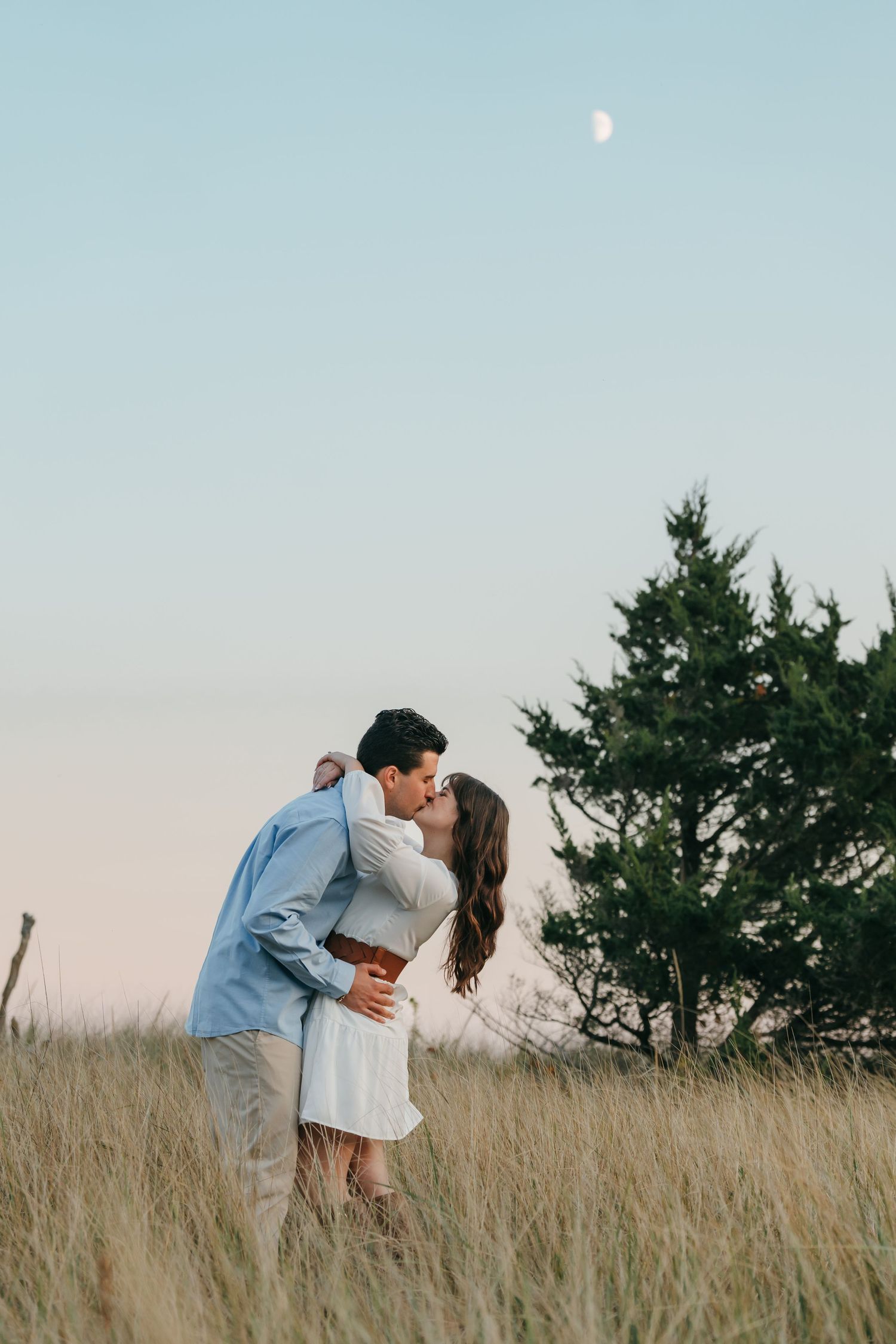 Romantic couple embraces in a grassy field at sunset with trees in the background.