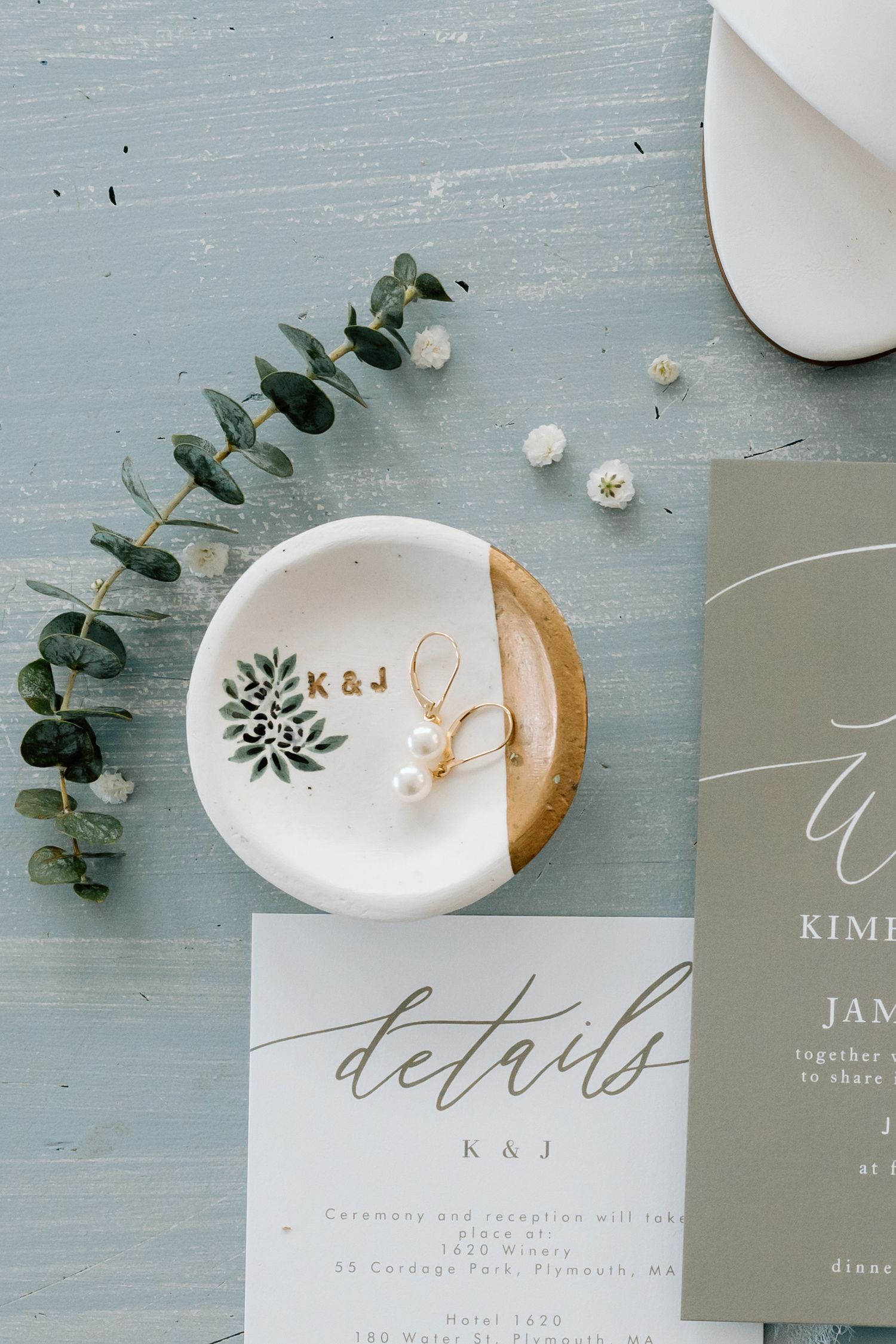 Wedding details flatlay with eucalyptus, ring dish, and invitation card on blue-gray surface.