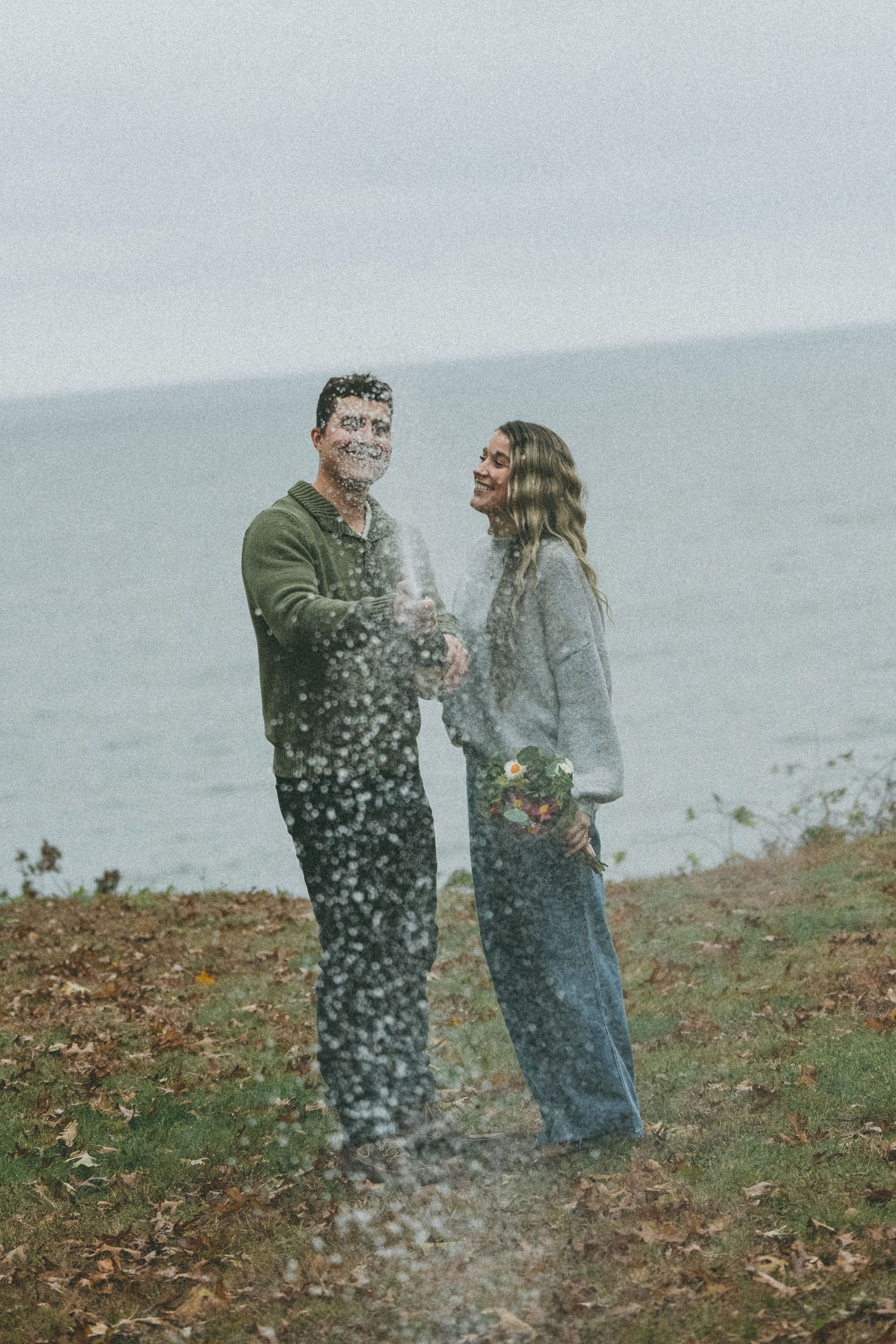 A couple laughs together as snow falls around them on a coastal hillside.