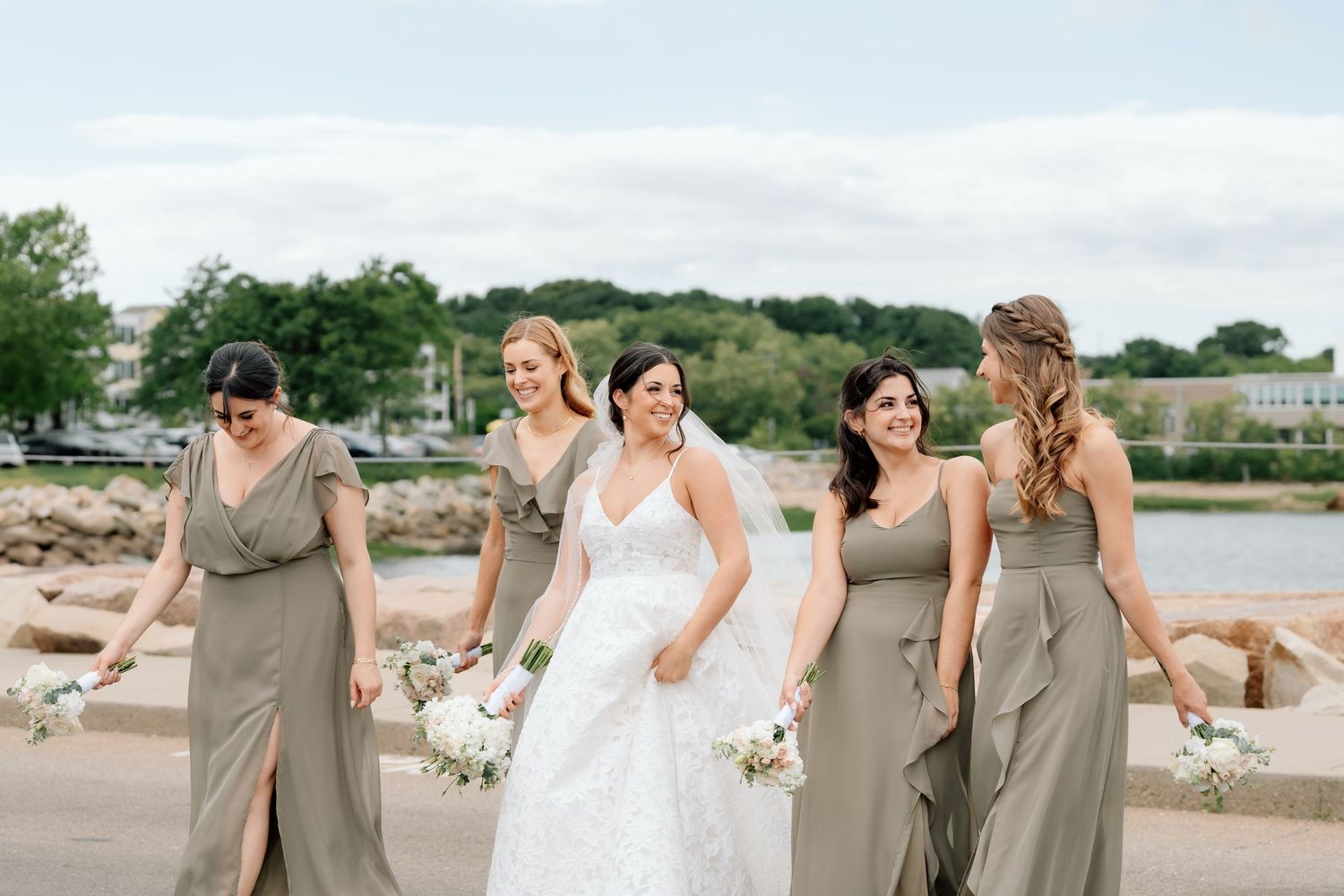 Wedding party in sage green dresses posing by waterfront with bouquets.