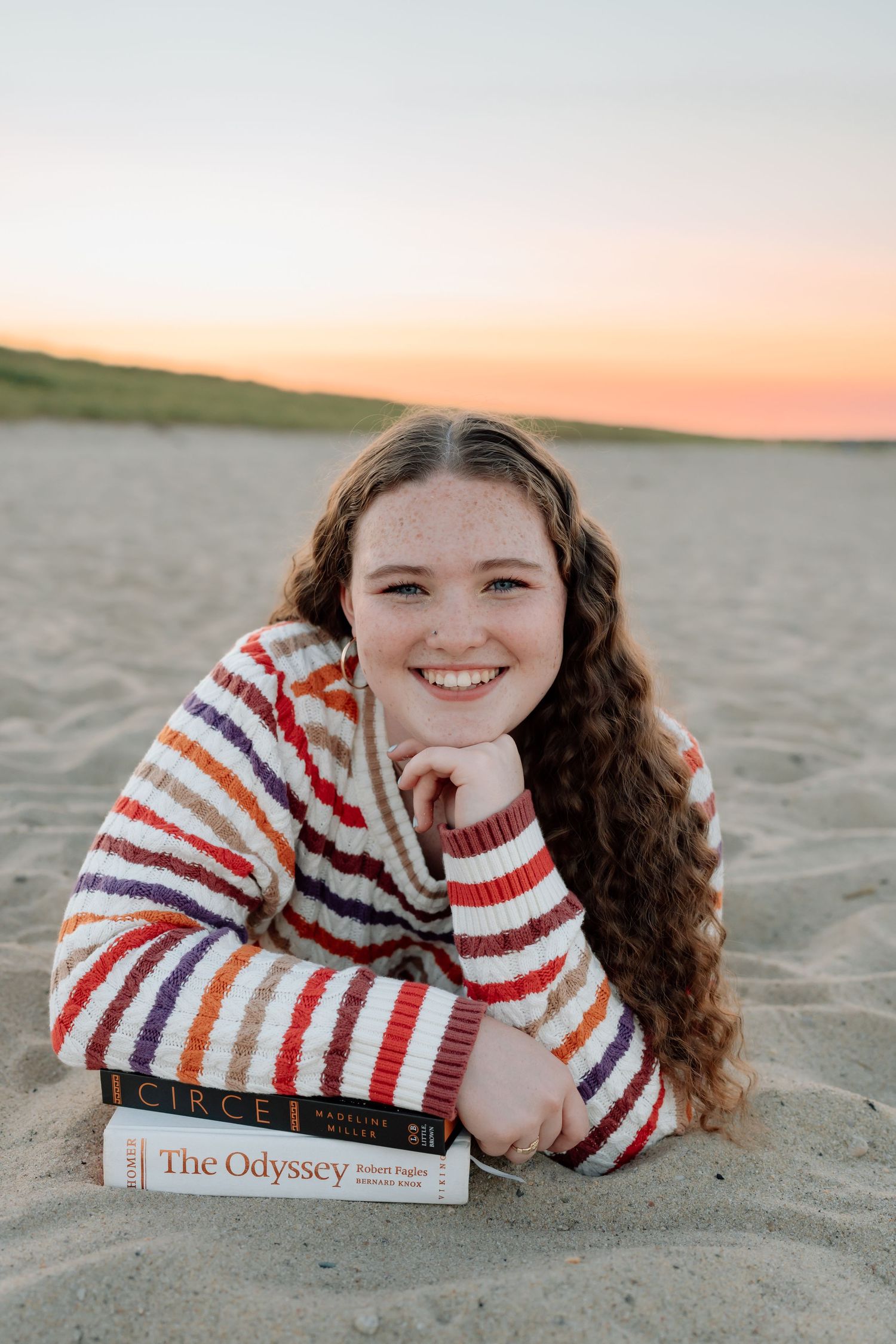Someone lying on beach sand at sunset with The Odyssey book and wearing colorful striped sweater.