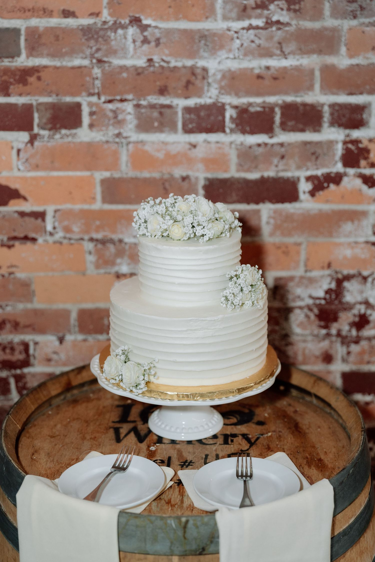 Simple white wedding cake displayed on wooden stand against rustic brick wall.
