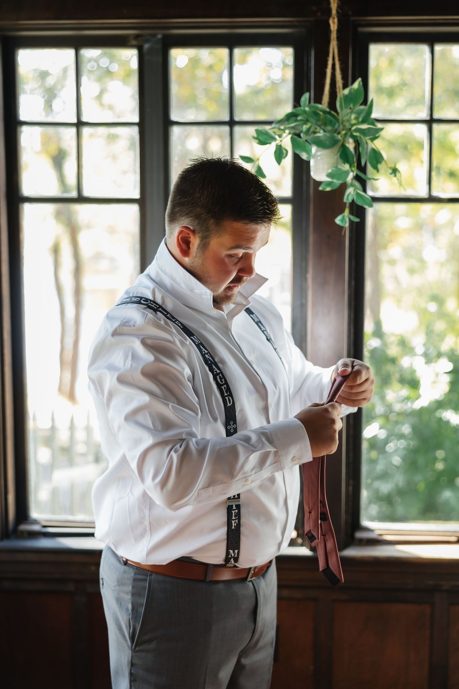A person in a white dress shirt and suspenders adjusts their tie while standing near a window.