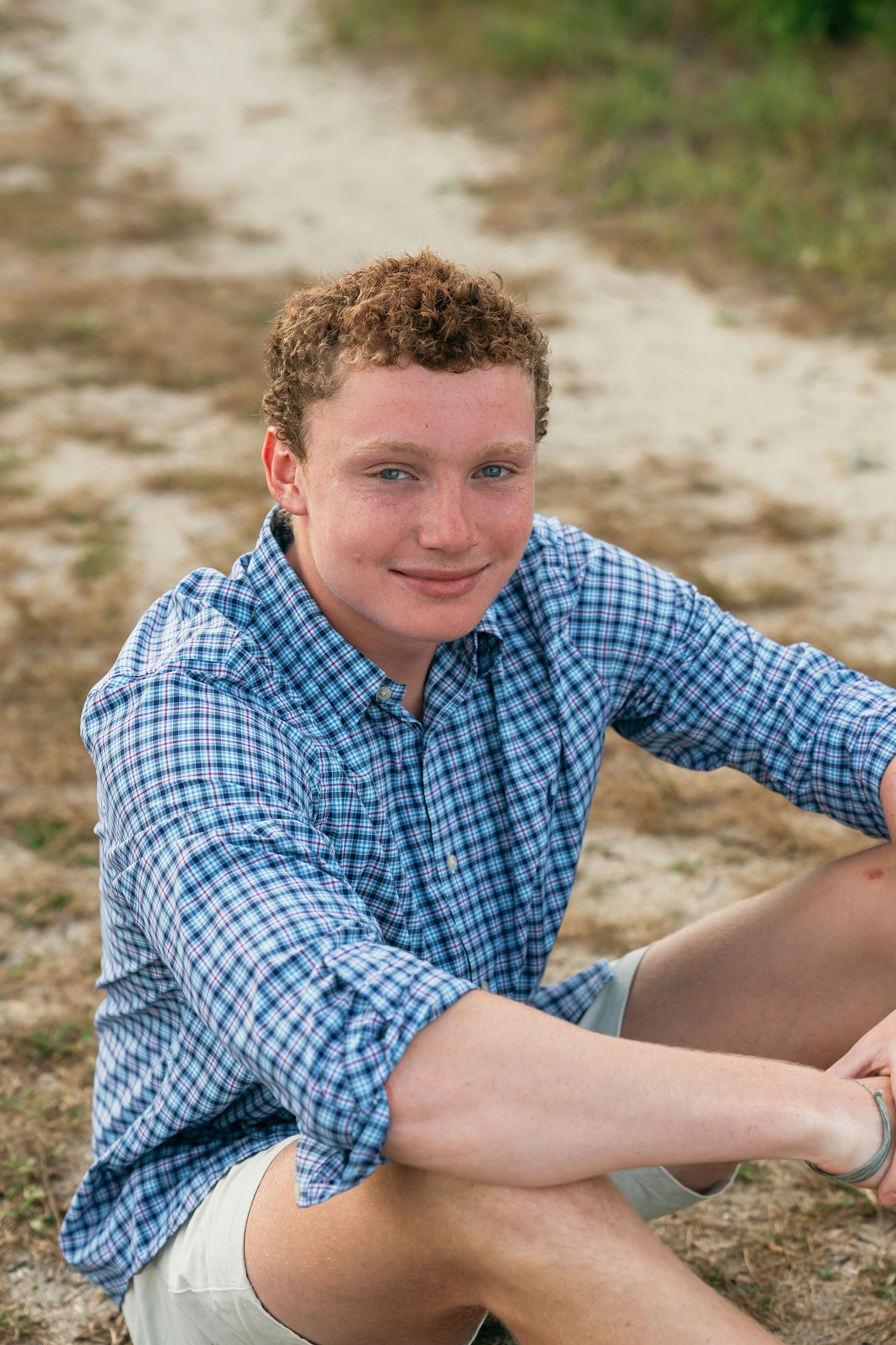 Person in blue plaid shirt sits casually on ground during outdoor portrait session.