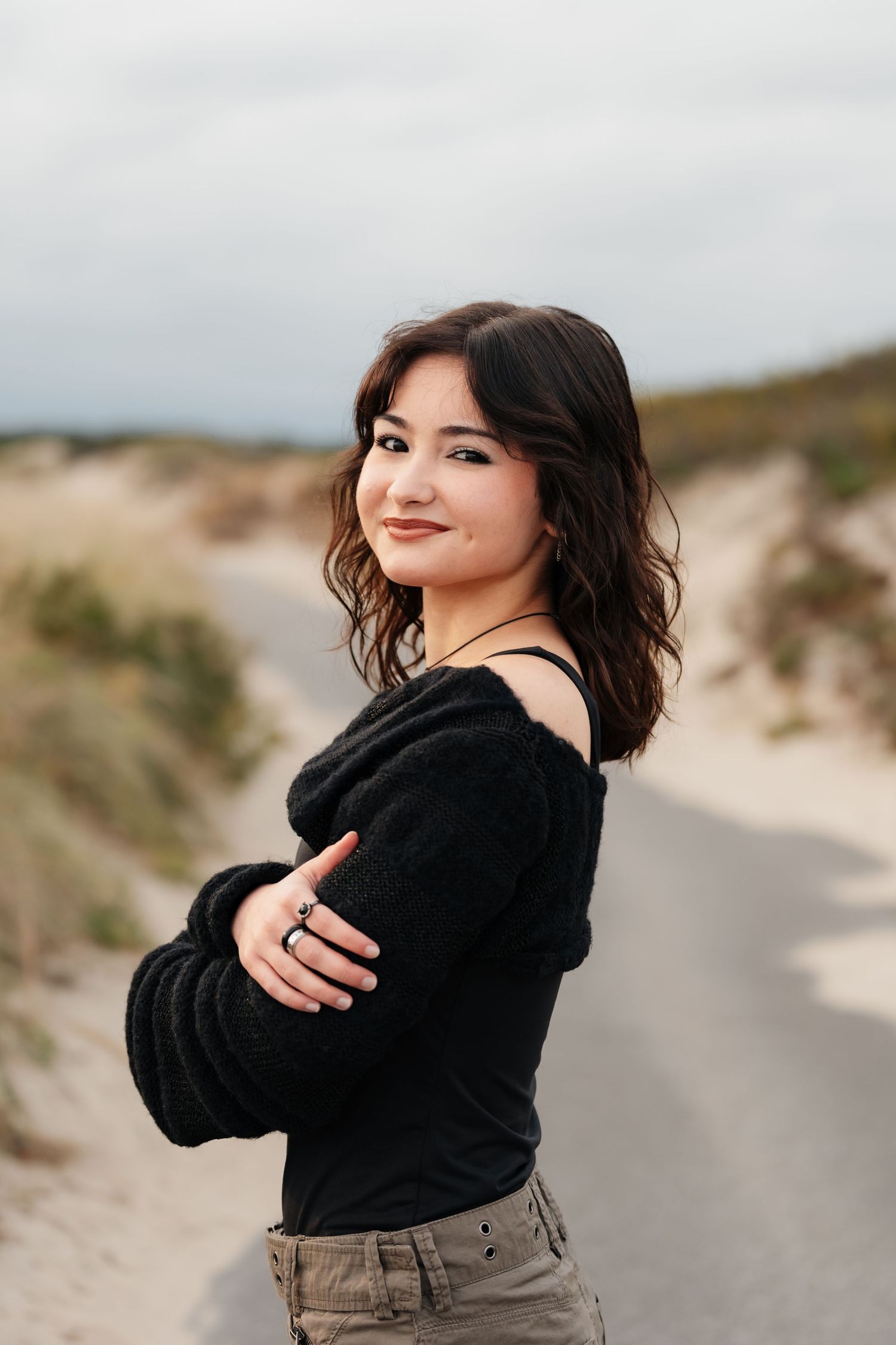 Person in black sweater poses against sandy dunes on a cloudy day.