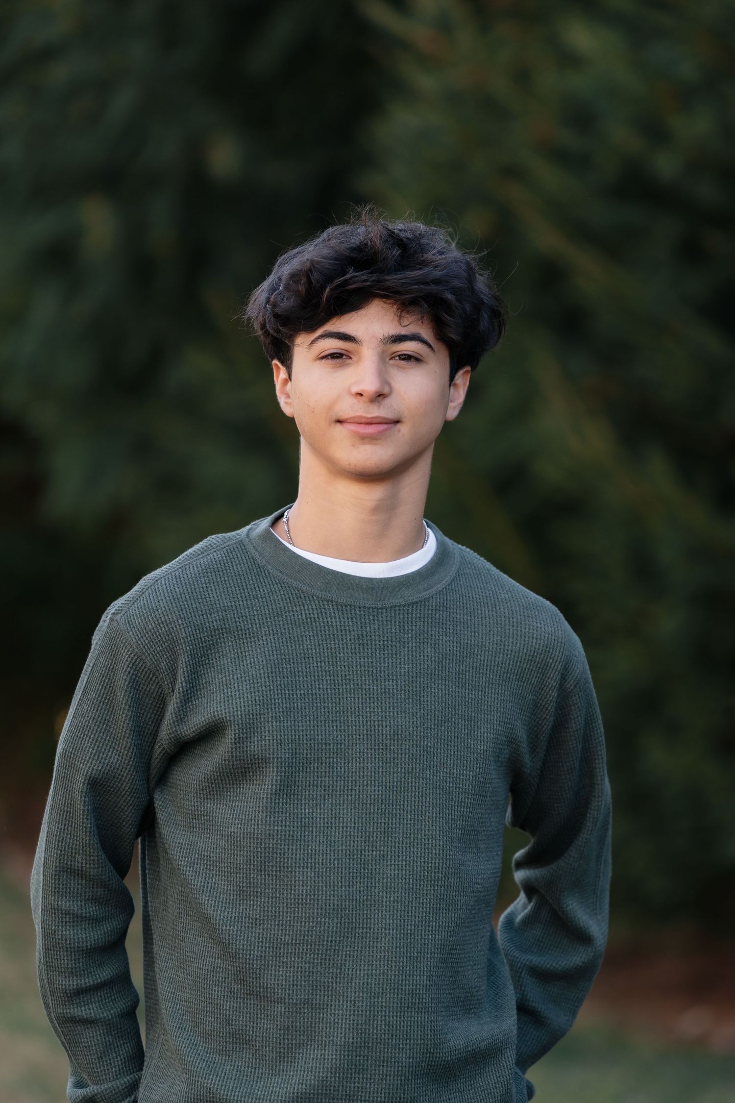 Person in green sweater poses outdoors near evergreen trees.