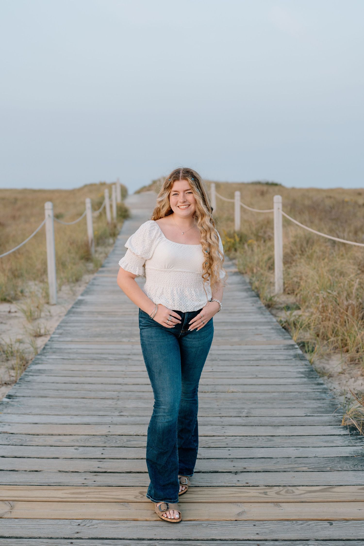 Person in white top walking on wooden boardwalk through coastal dunes.