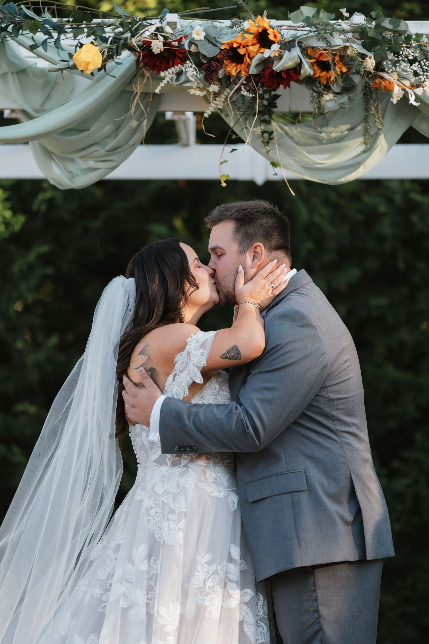 Newlyweds share their first kiss under an arbor decorated with autumn flowers.