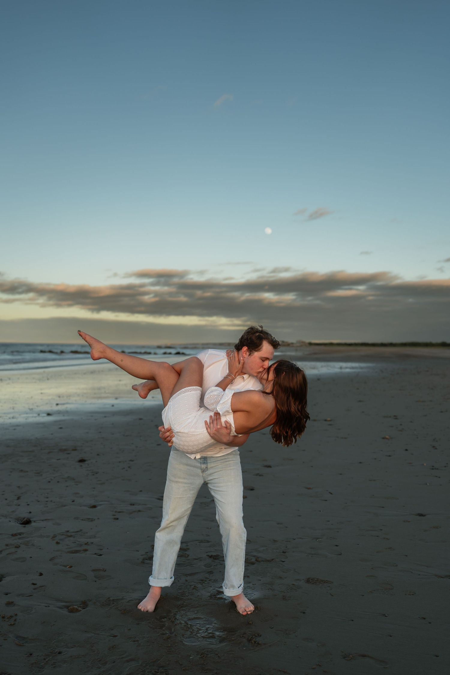 A couple performs a romantic dip pose on a beach at sunset.