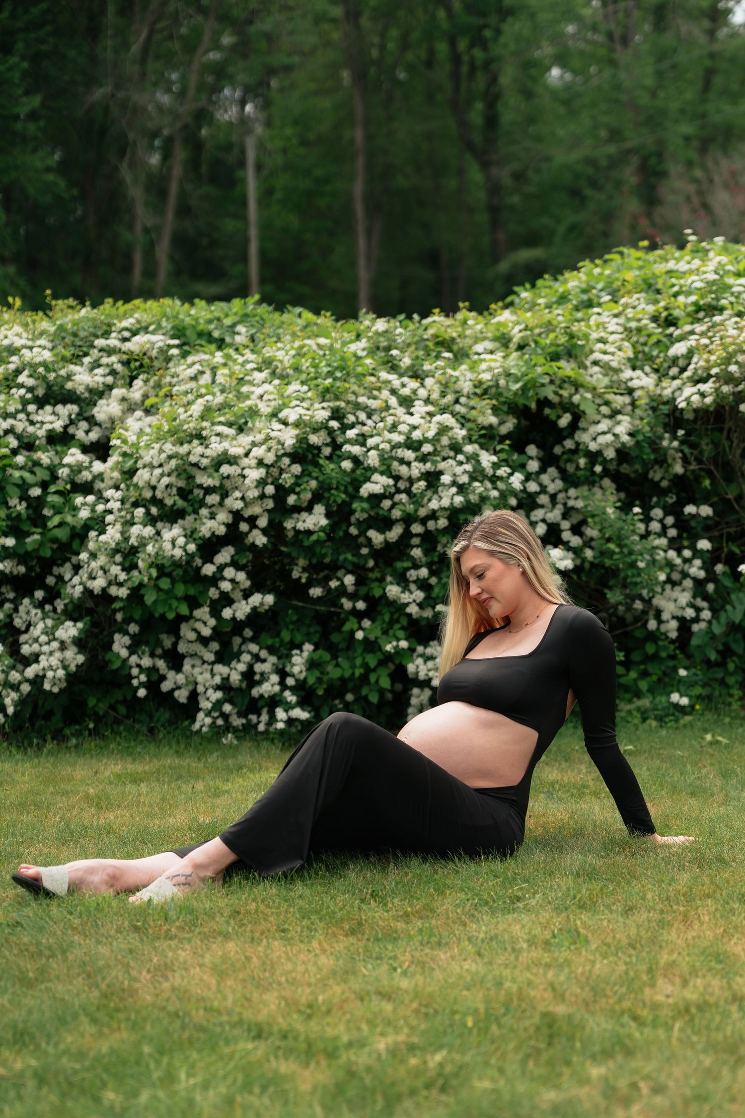 A pregnant woman in a black dress reclines gracefully on green grass near blooming white flowers in a garden setting.