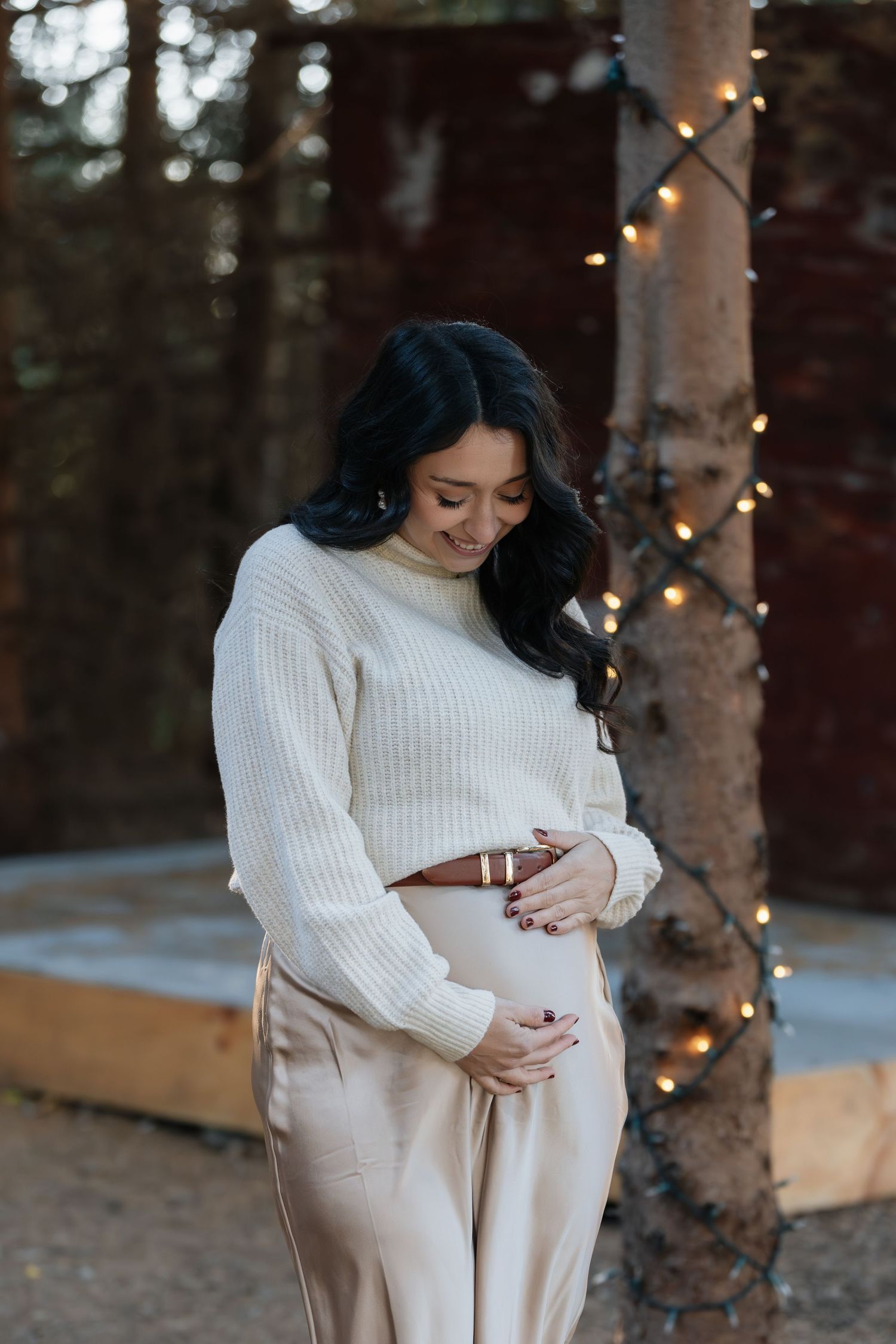 Maternity photos in white sweater and beige pants among trees decorated with string lights.