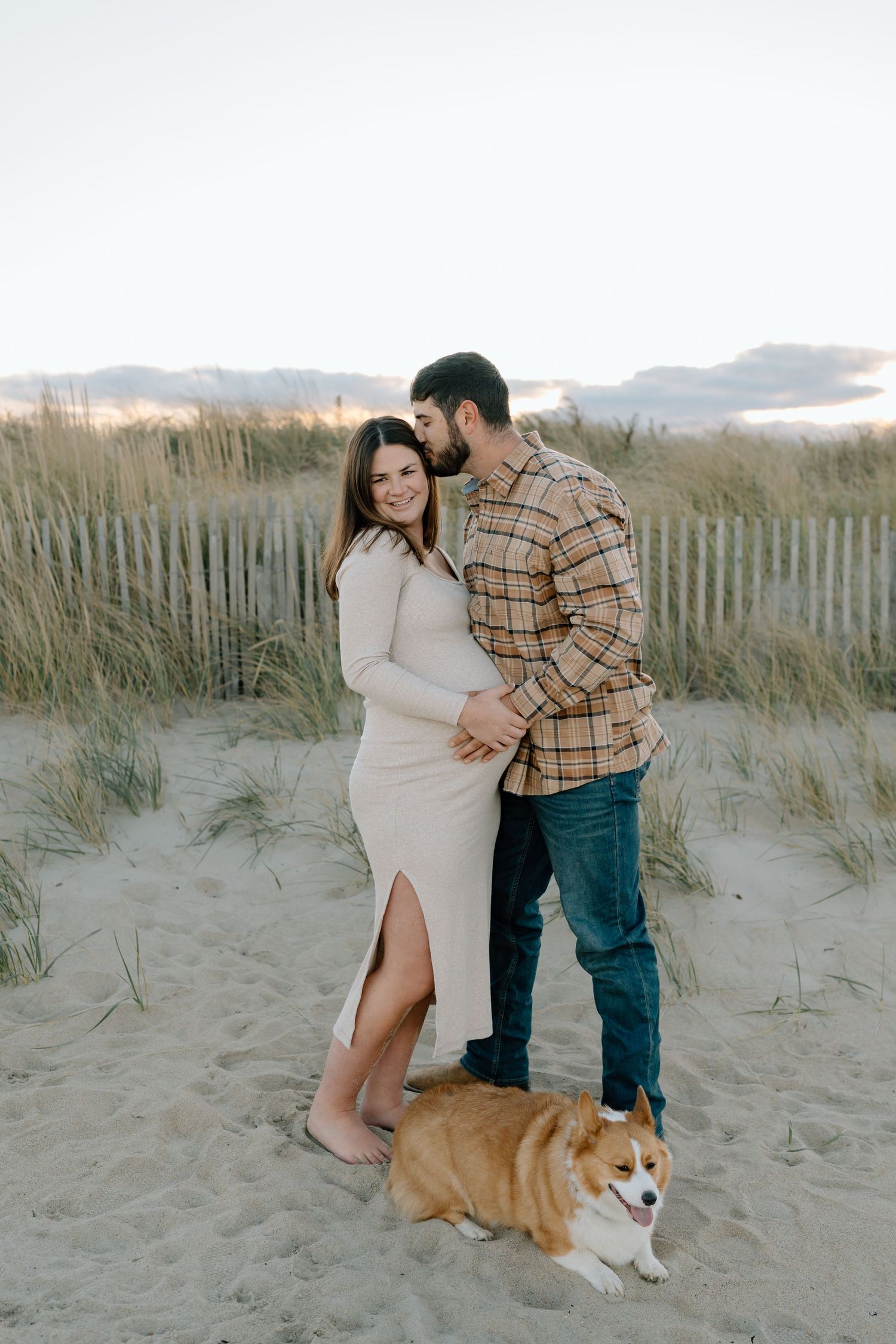A couple poses with their Corgi on sandy beach dunes during sunset.