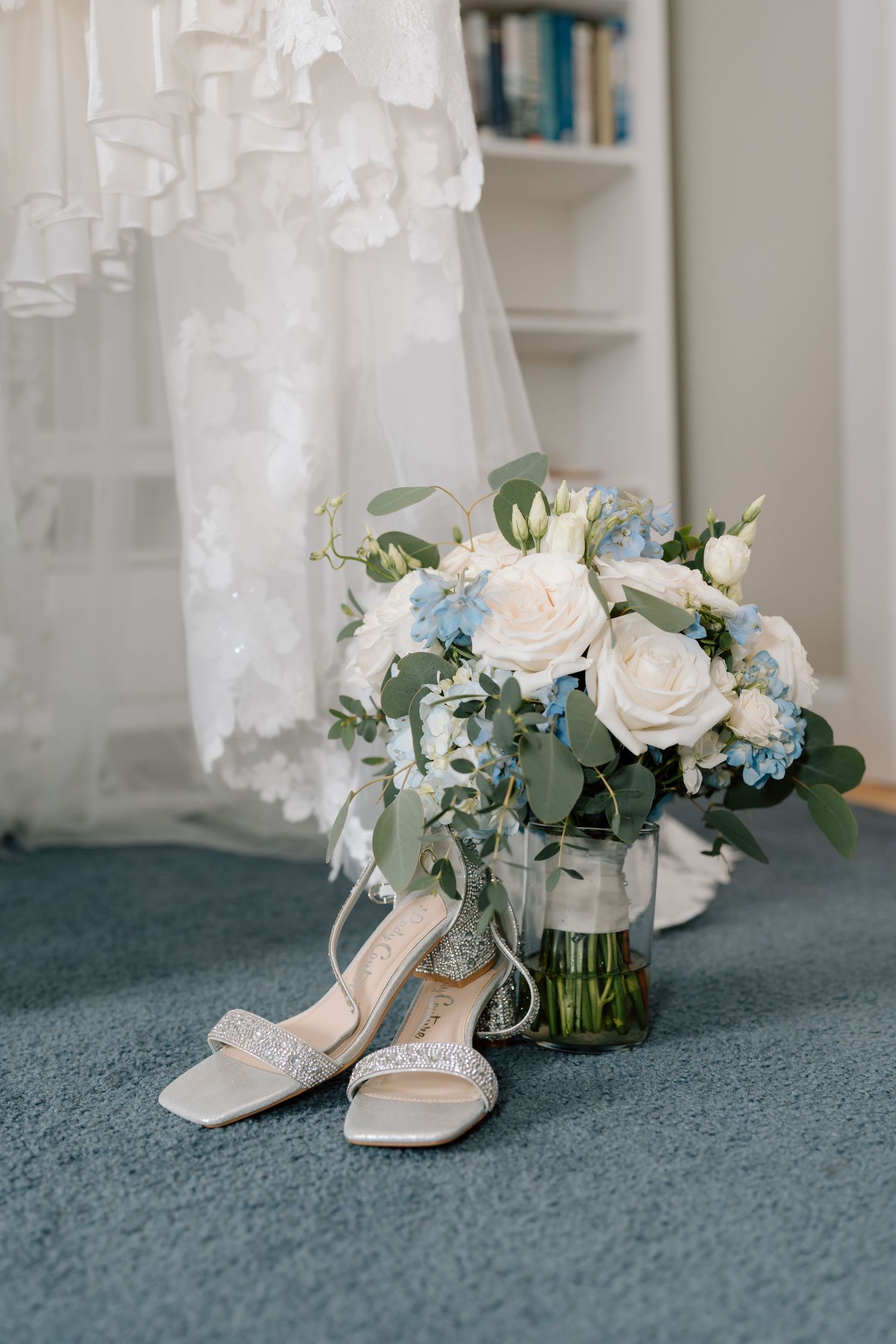 Bridal shoes and bouquet with white roses and blue flowers arranged on blue carpet.