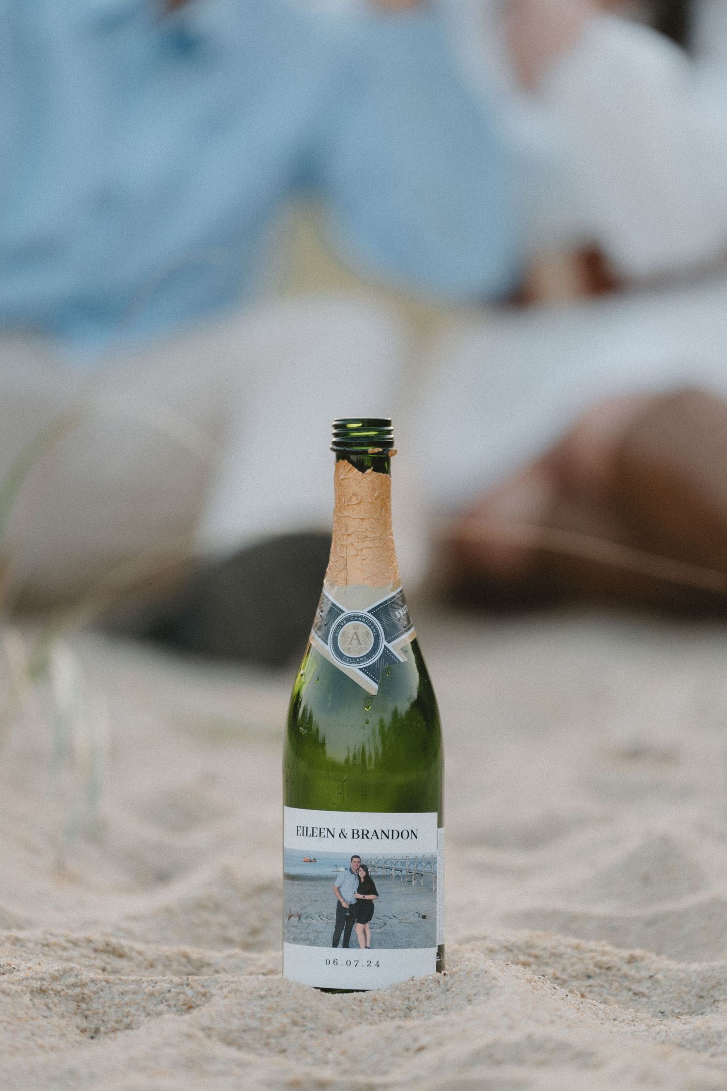 A personalized champagne bottle with a custom label sits in the sand at sunset.
