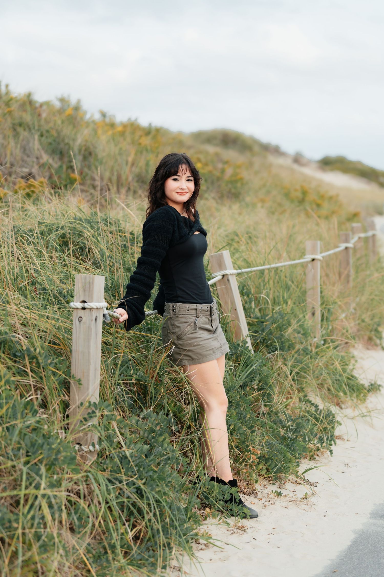 Person in dark outfit poses along sandy coastal path with fence.