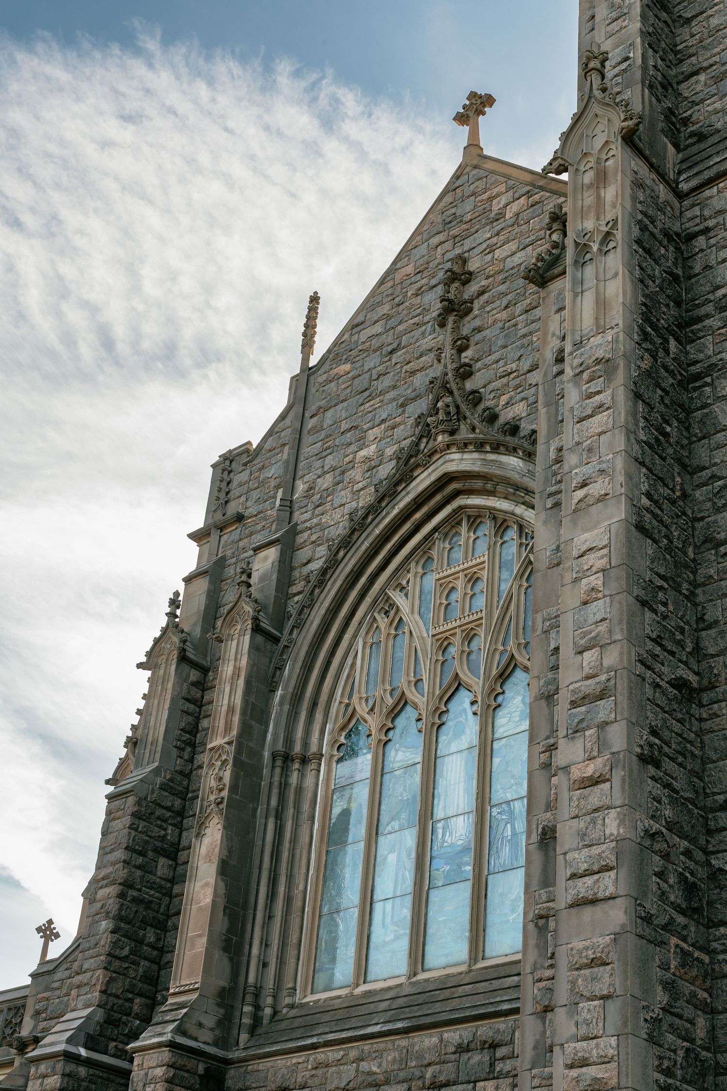 Gothic stone church exterior with arched stained glass windows and ornate architectural details against cloudy sky.